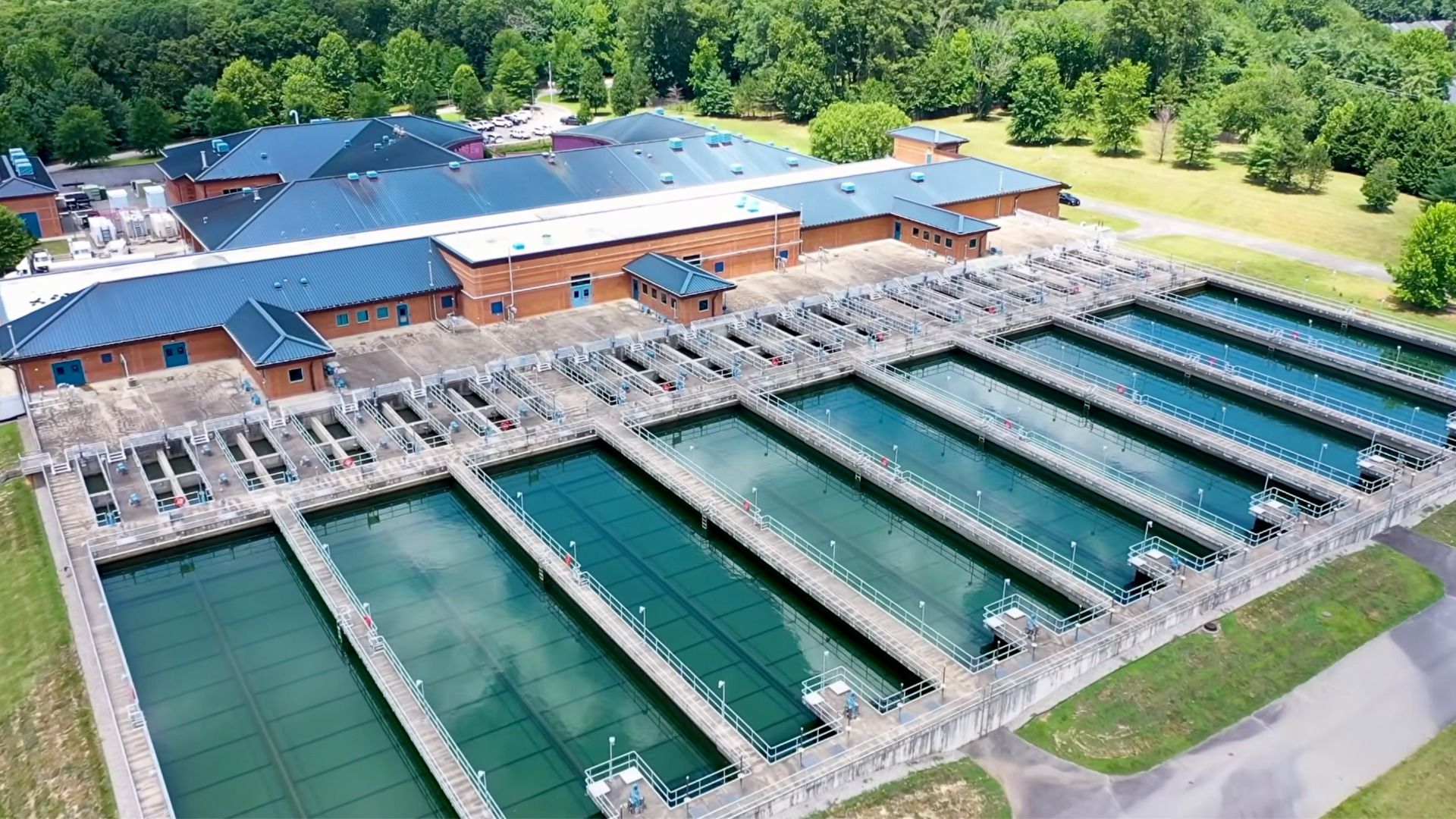 Aerial view of a wastewater treatment plant with long rectangular basins of teal water, metal walkways, and brick buildings with blue roofs, set among green trees and grass.