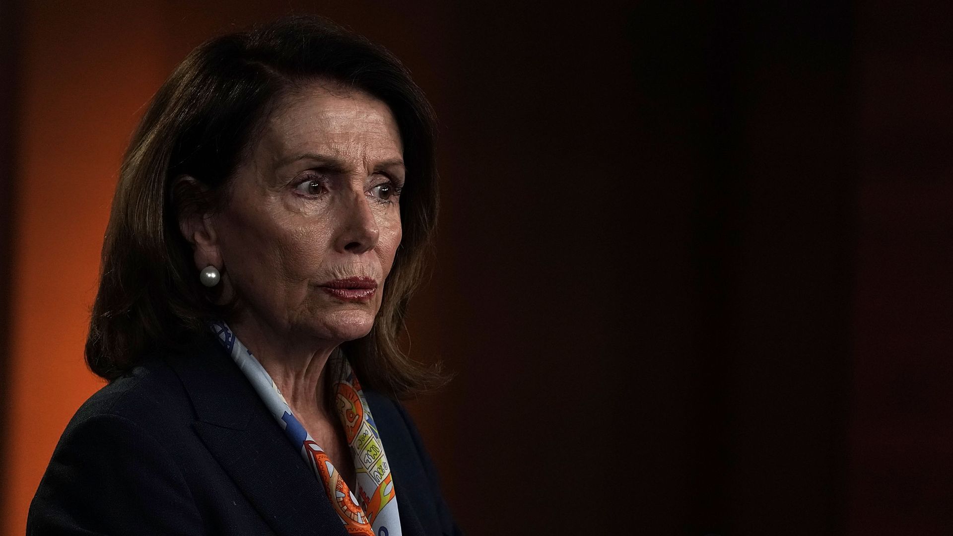 Nancy Pelosi in a dark suit with a colorful scarf before a black and orange backdrop.