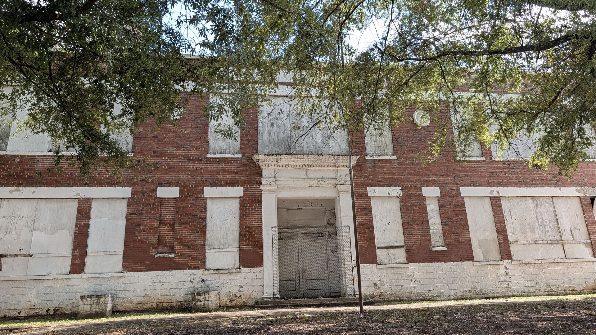 Abandoned red-brick building with boarded windows, a central stone-framed doorway, white basework, and a chain-link gate; overhanging tree branches obscure the facade.
