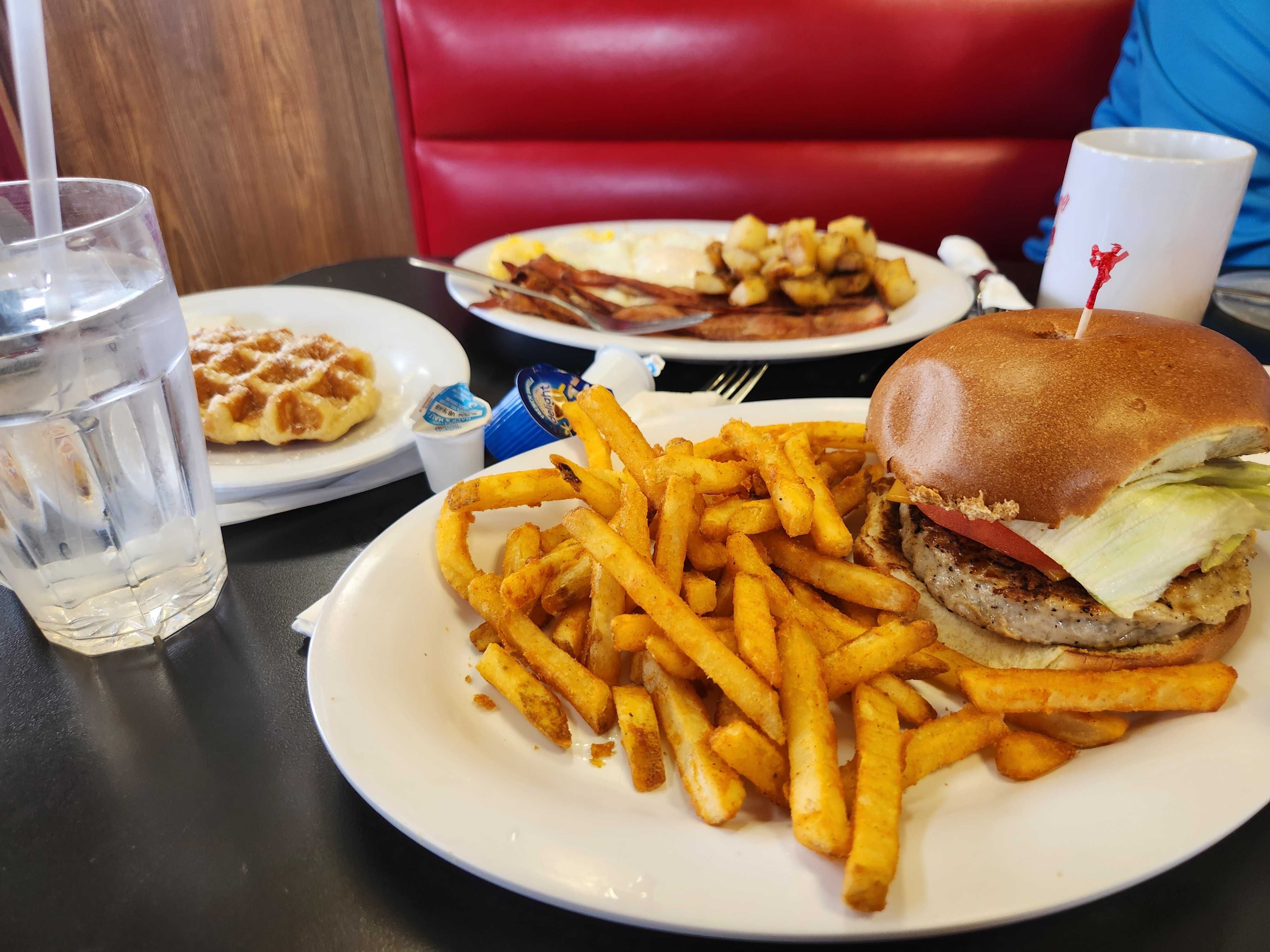 Burger with lettuce and tomato and a large plate of fries in the foreground; background shows a bacon-and-eggs plate with potatoes. A glass of water on the left and a white mug on the right.