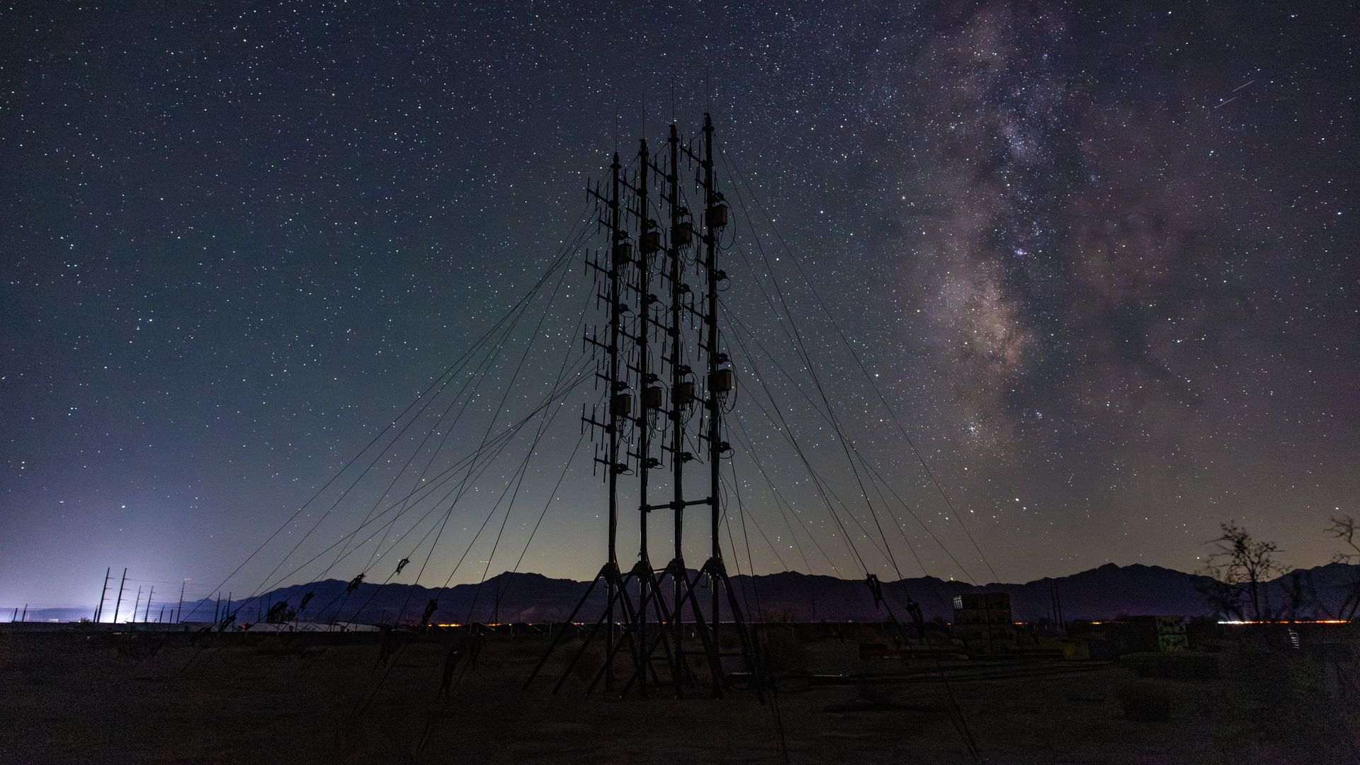 Night sky filled with stars and the Milky Way over a silhouetted tall metal structure with multiple antennas, cables, and distant mountain range in the background.