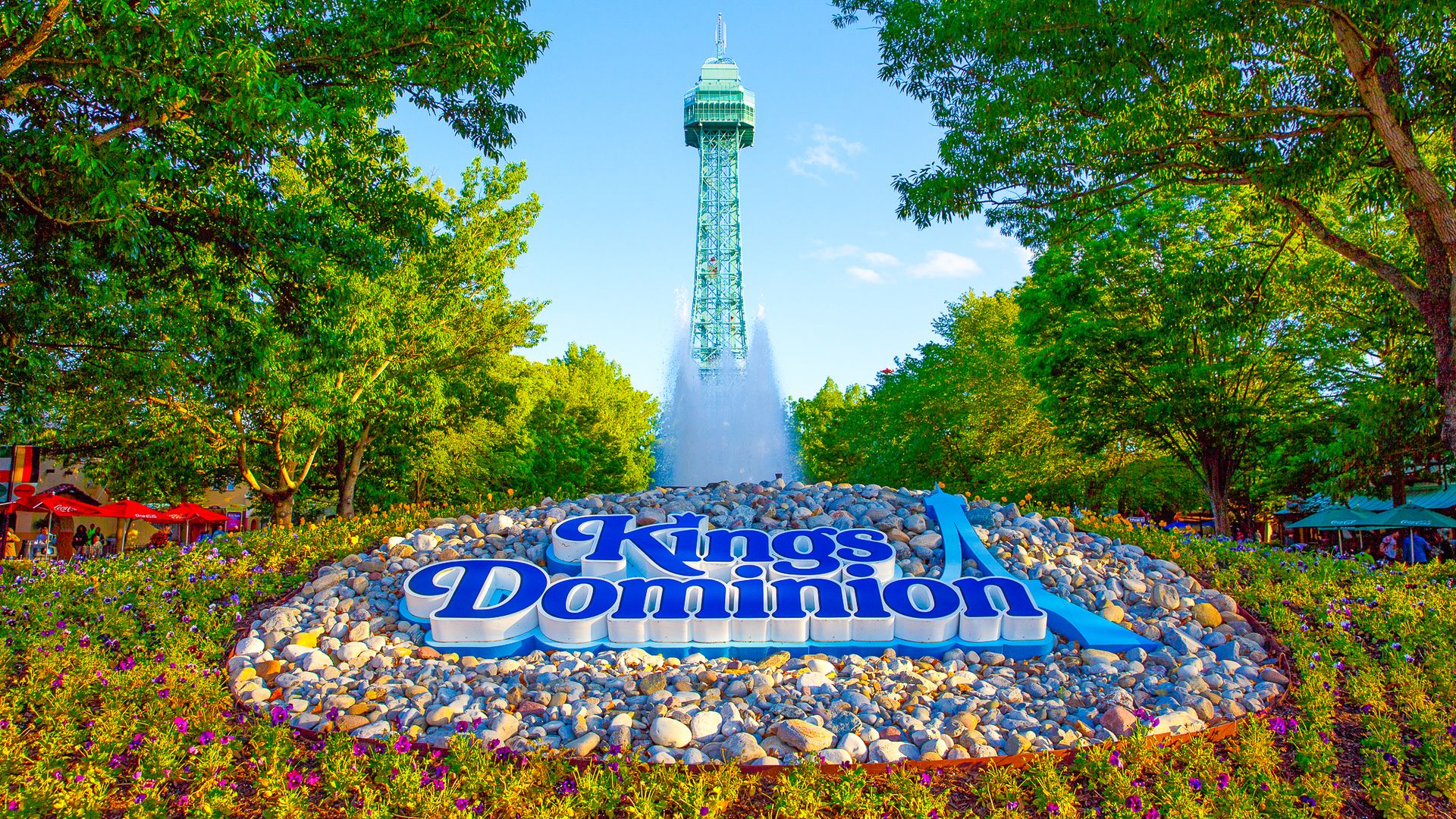 Bright day at Kings Dominion: a tall blue-green tower rises behind a water fountain, framed by lush trees. A large blue and white Kings Dominion sign sits on a bed of rocks with a flower border.