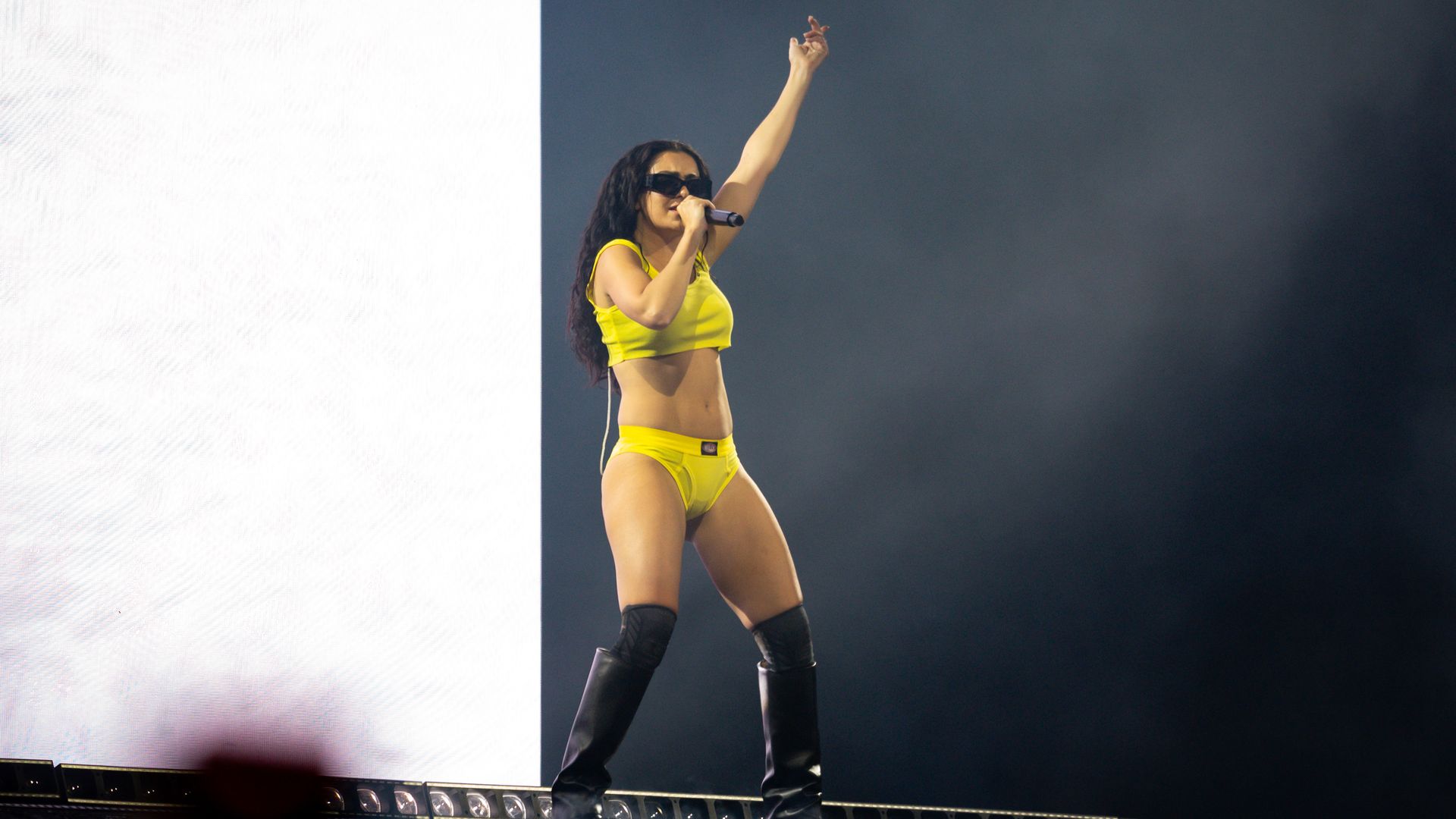 Female singer in a bright yellow crop top and shorts, black sunglasses, knee-high boots, microphone in hand, raises one arm on stage against a dark backdrop with a large white screen on the left.
