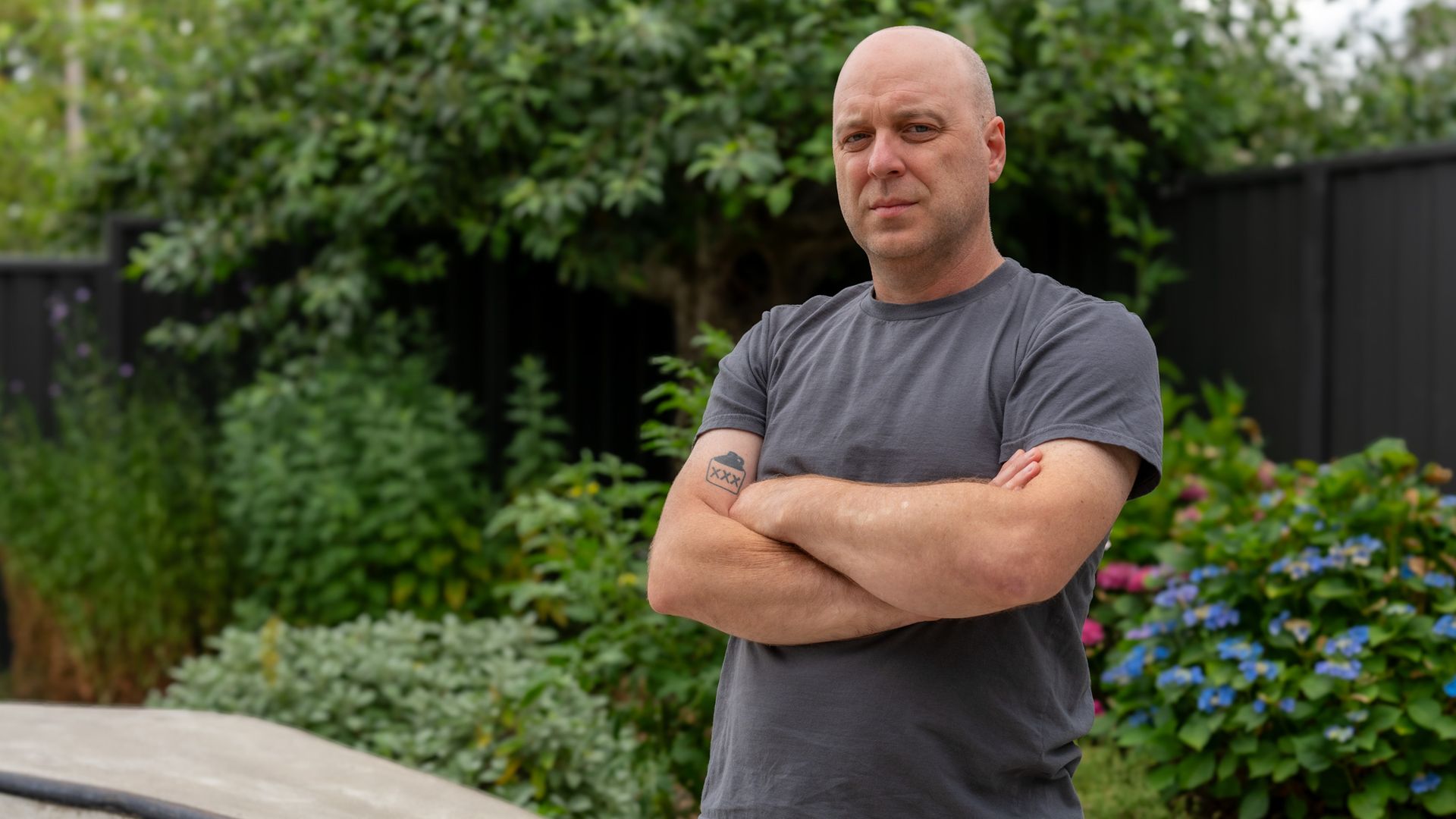 Bald white guy arms folded in a grey t-shirt standing in front of bushes and his concrete skateboard ramp