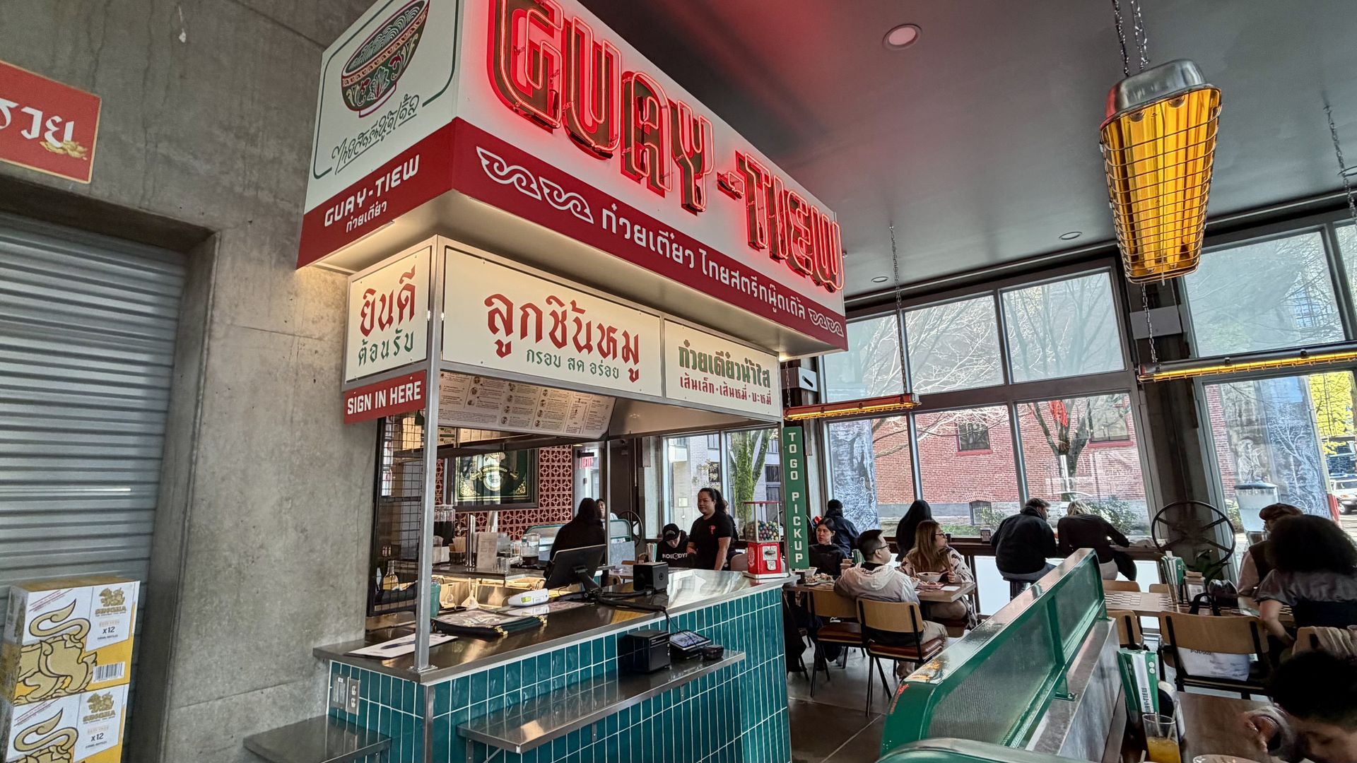 Thai noodle shop interior with a turquoise tiled counter, a red neon sign reading "GUAY-TIEW" above, Thai menu boards, and customers seated by large windows; warm hanging heaters and natural daylight.