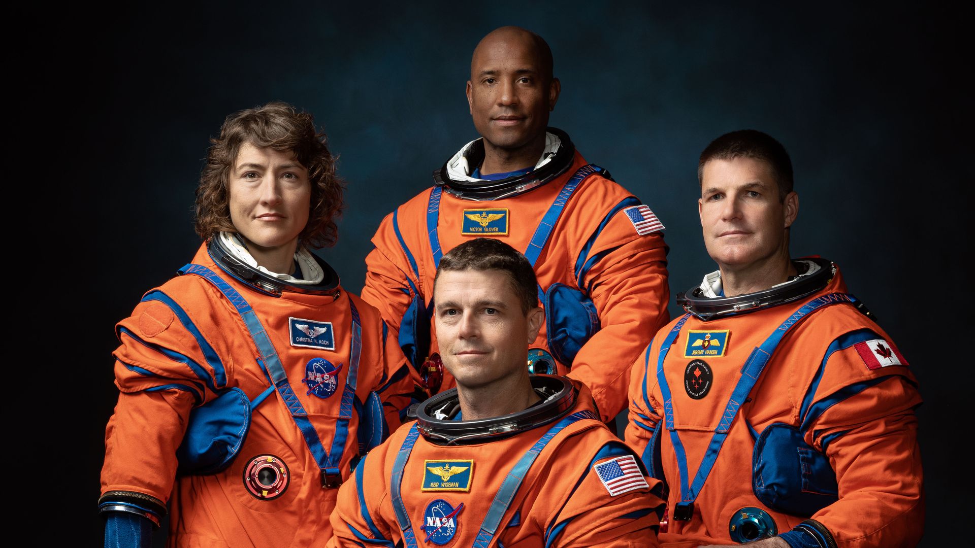 Four astronauts pose in their orange spacesuits with helmets off