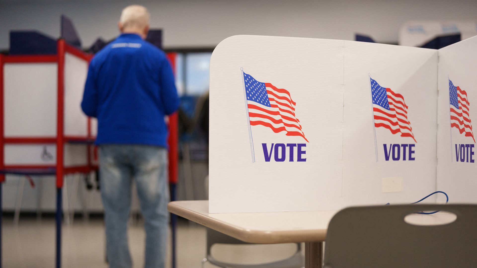 A polling station with white voting booths displaying a waving American flag and the word VOTE; a man in a blue jacket stands near red voting booths in the background.