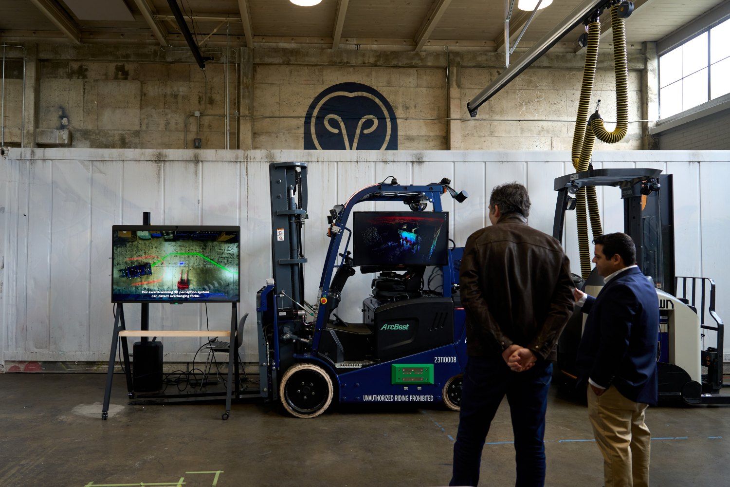 Two men observe a smart forklift system in a warehouse, with screens showing real-time navigation data and the Samsara logo in the background.