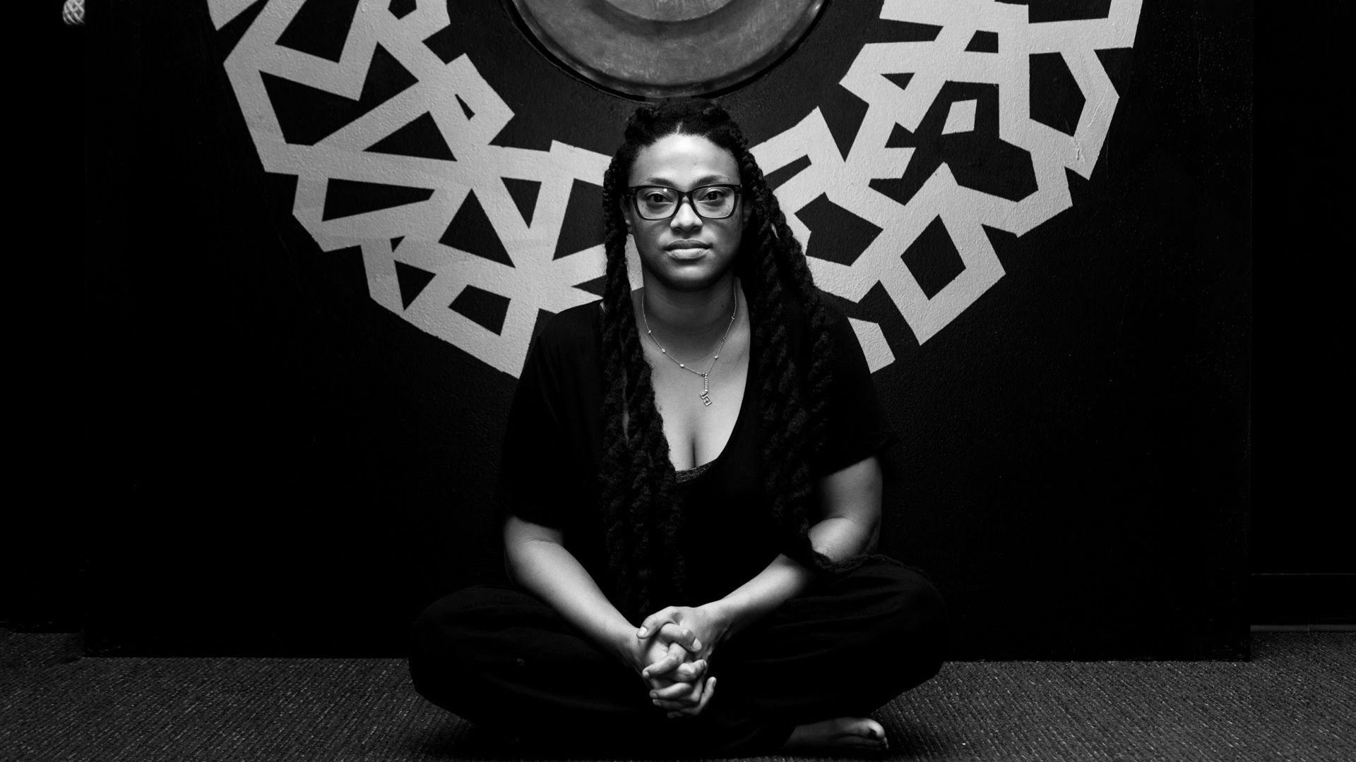 A black-and-white photo of artist Ya La'Ford sitting on the ground cross-legged.