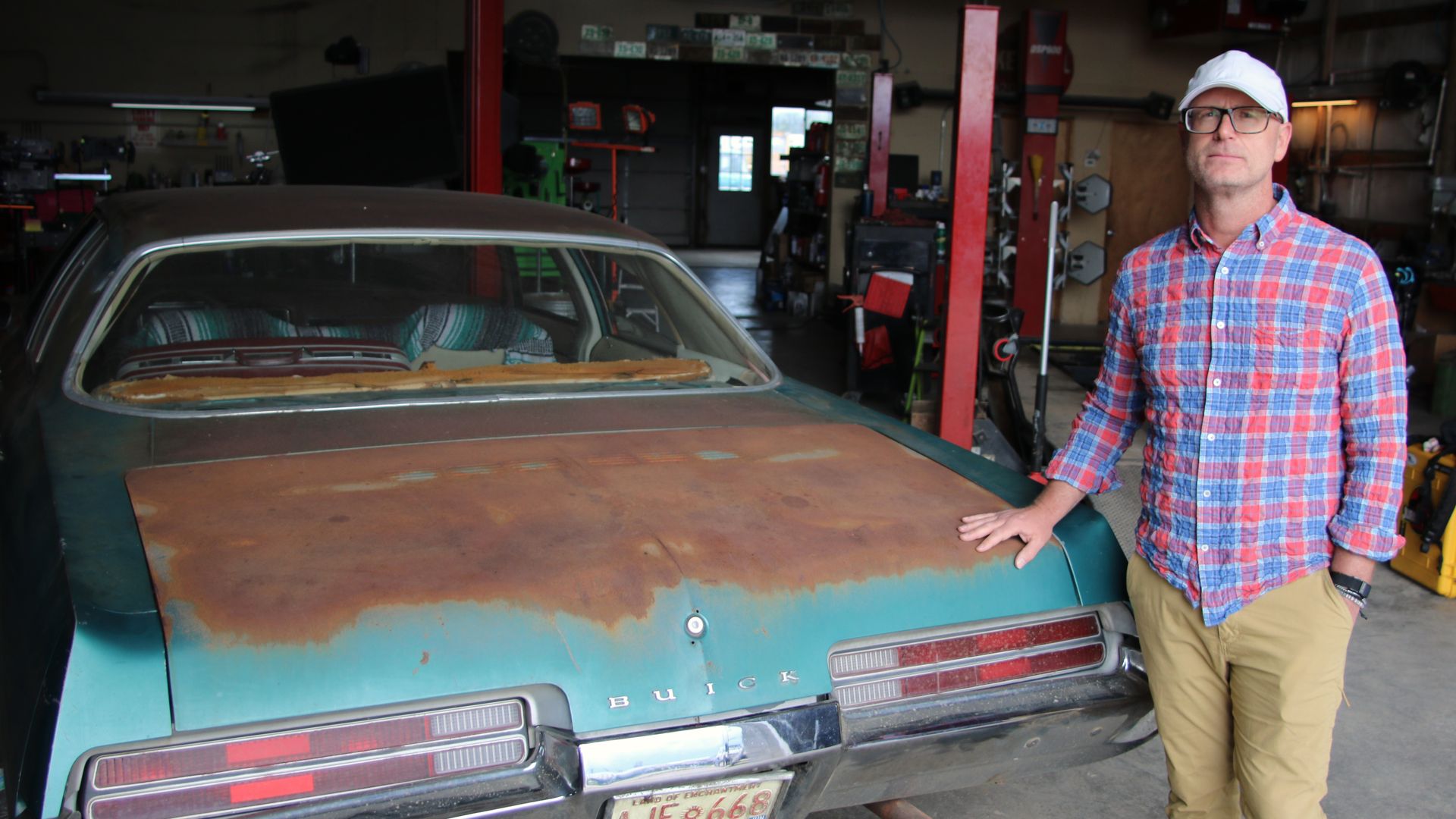 A man in a plaid shirt and white cap stands beside a rusted turquoise 1971 Buick LeSabre in a cluttered auto shop, with tools and equipment visible in the background.