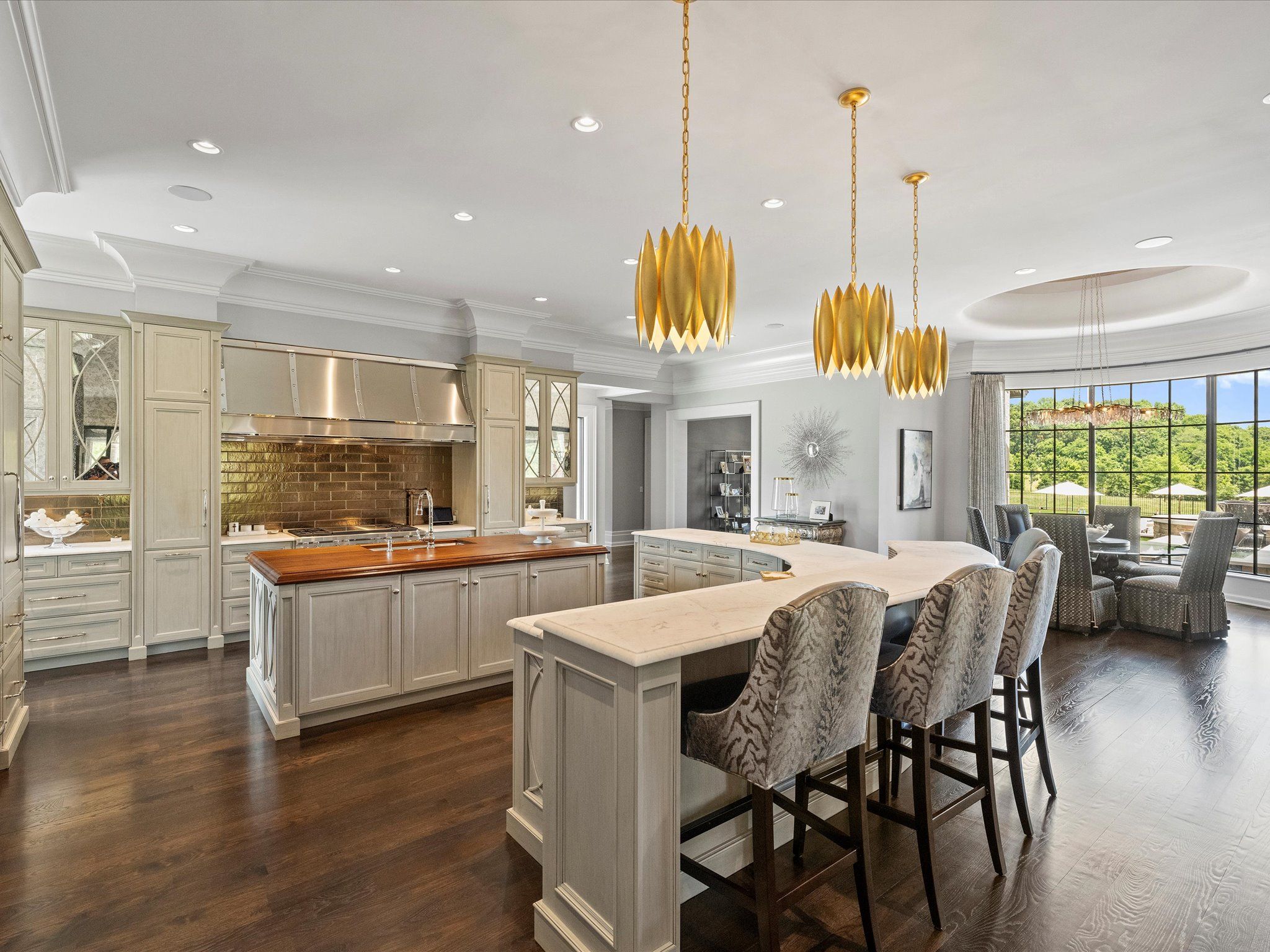 Spacious modern kitchen with cream cabinets, dark wood floors, marble and wood countertops, three gold pendant lights, bar stools with patterned upholstery, large windows and dining area with gray chairs.