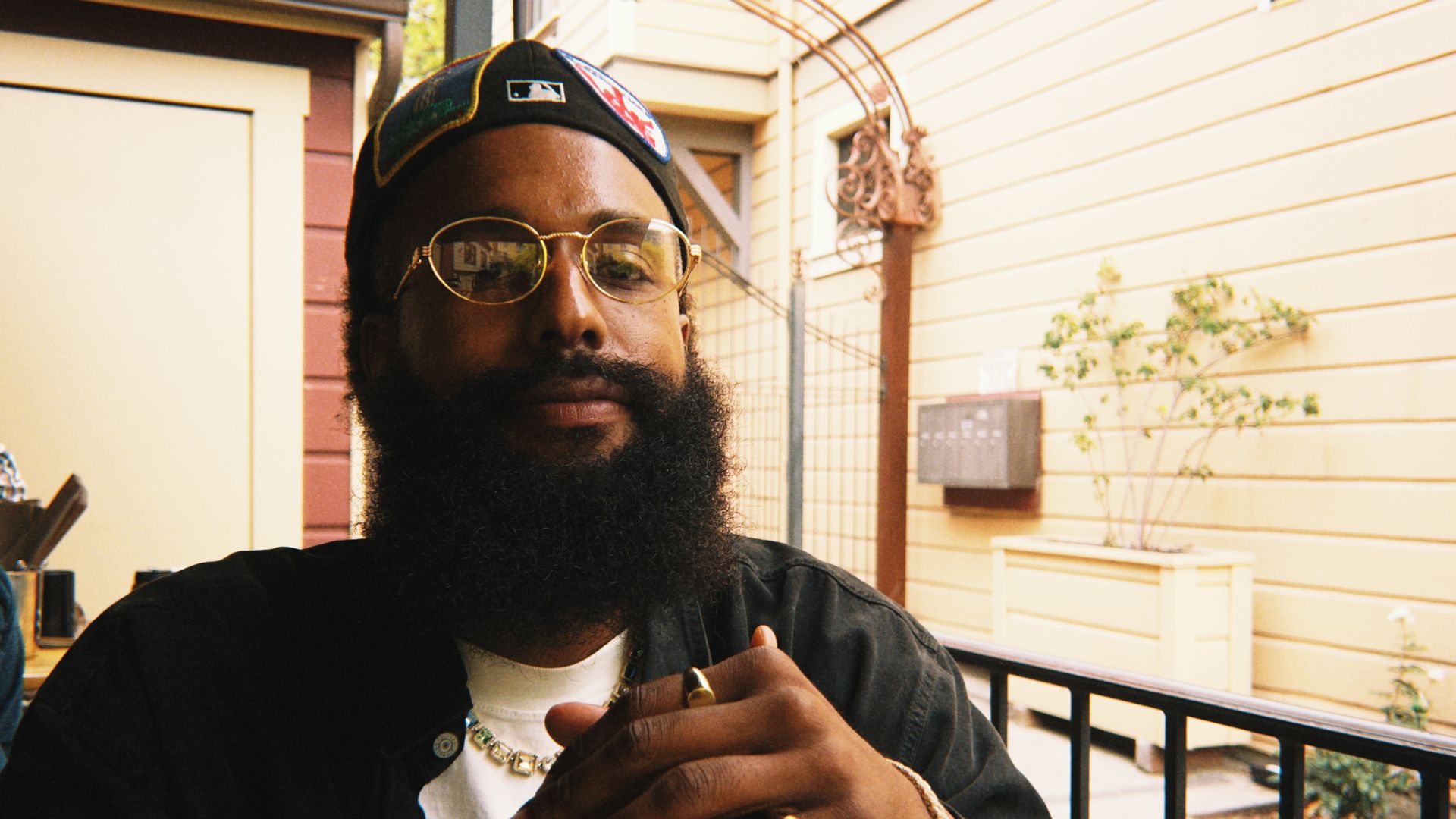 Man with a thick black beard and glasses wearing a black cap and jacket, seated outdoors with hands clasped, gold rings and bracelet visible, light-colored building in background.