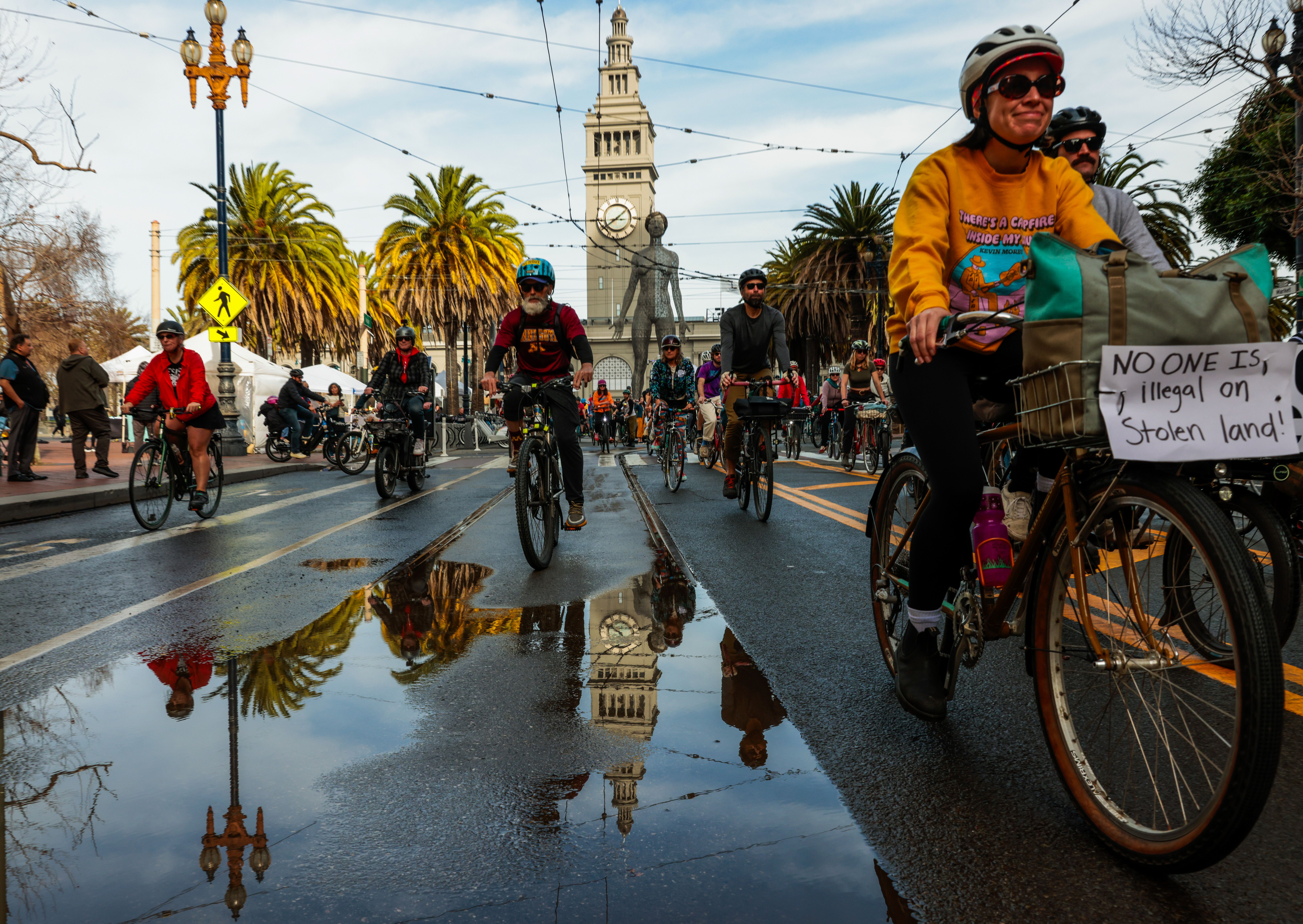 People riding bicycles on a street with a clock tower in the background. One rider is wearing a University of Minnesota shirt and another has a sign that says "no one is illegal on stolen land"