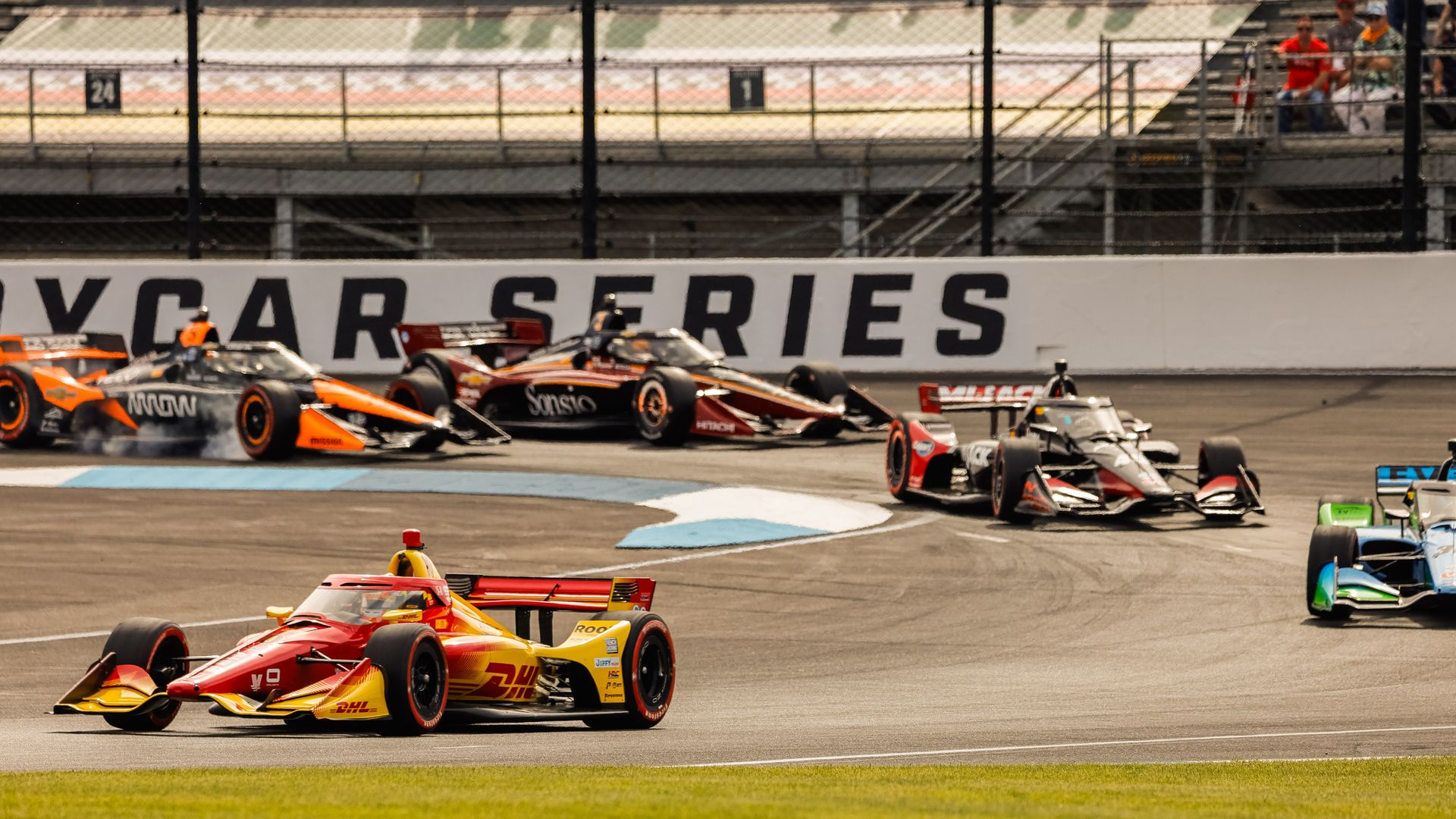 Wide shot of a Formula race on a track: a bright yellow DHL car leads orange and black racers. A white wall reads CAR SERIES as spectators watch from the grandstands behind a chain-link fence.