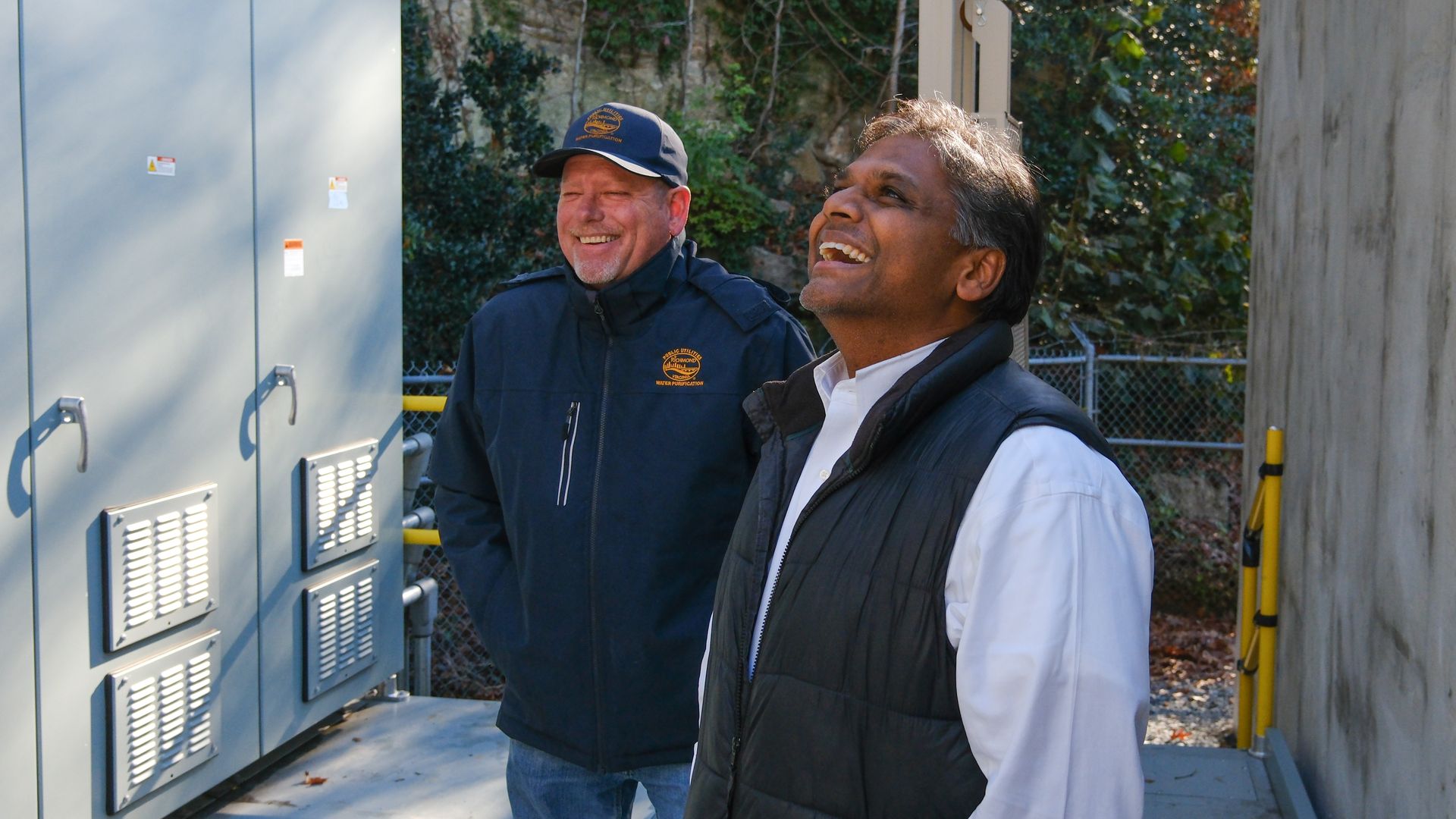 Two men outdoors near electrical panels, one in a blue jacket and cap, the other in a black vest over a white shirt, both smiling and looking up, with trees and a fence in the background.