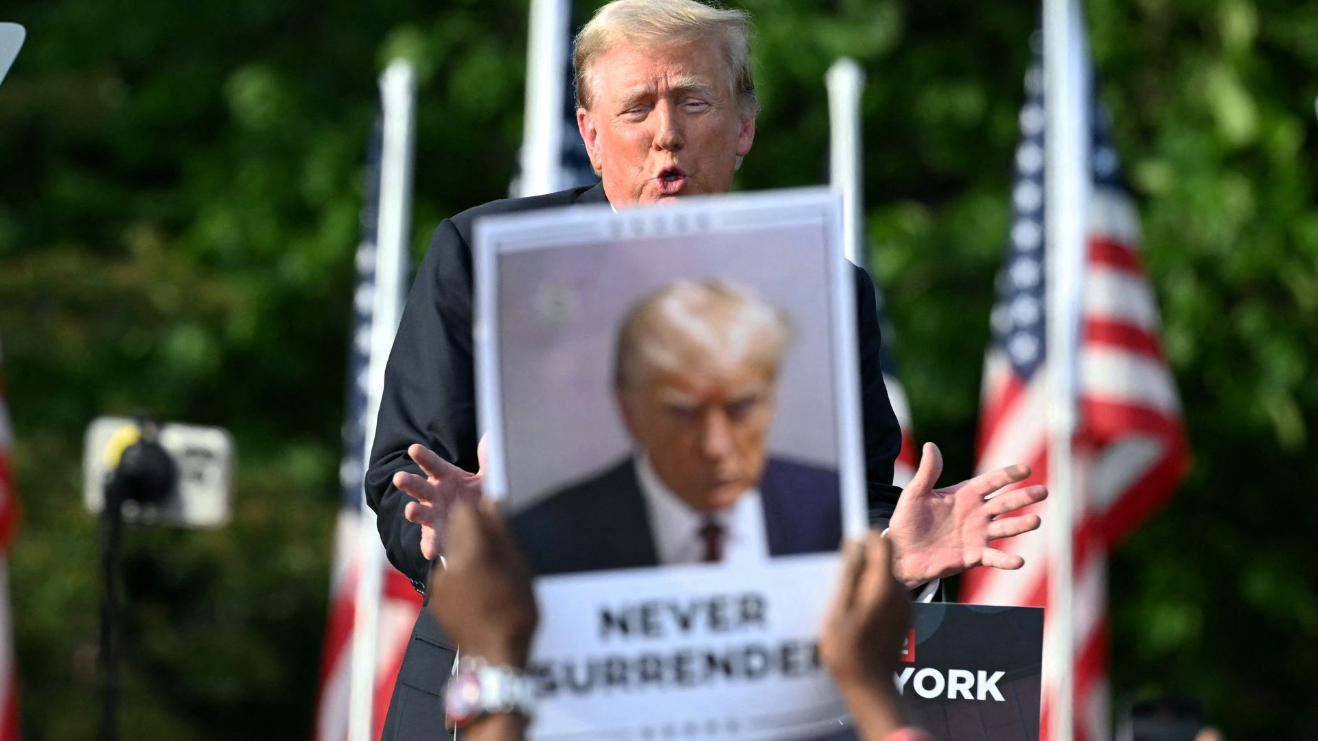 Trump speaks from a podium, partially obscured by a supporter's "Never surrender" sign.