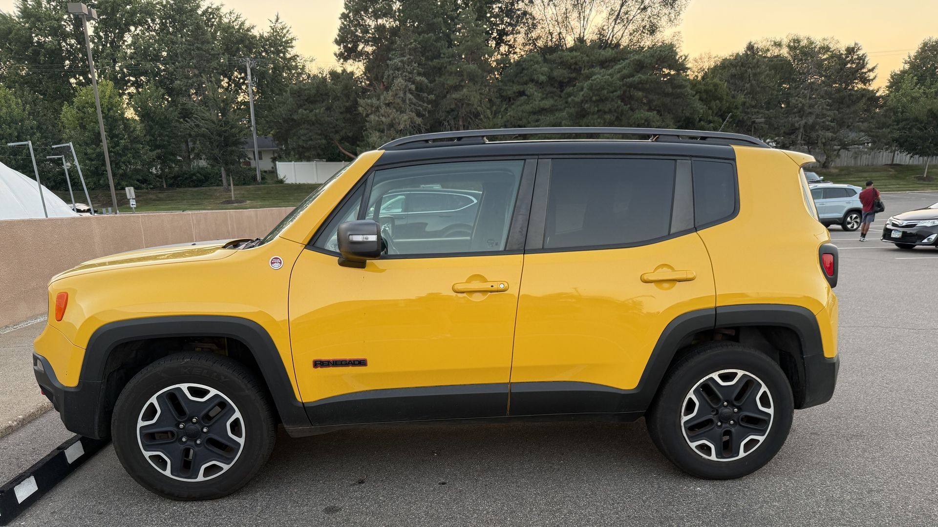 Side view of a bright yellow Jeep Renegade in a parking lot, roof rails visible, black trim and big tires. In the background, a person in a red shirt near a blue SUV; trees and a pale sunset sky.