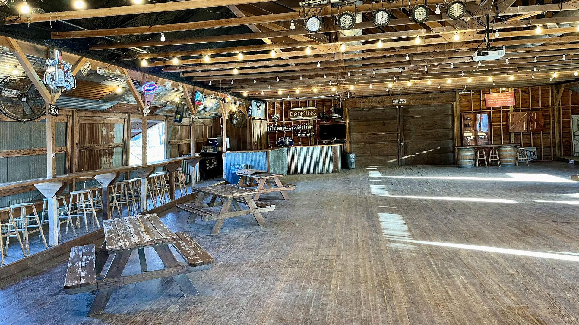 A rustic barn-style bar and hall with a wooden floor, long bar and stools on left, picnic tables, string lights on exposed beams, and a back area labeled "DANCING" under stage lights. This is the The Albert Dancehall in the Texas Hill Country, between Fredericksburg and Johnson City.