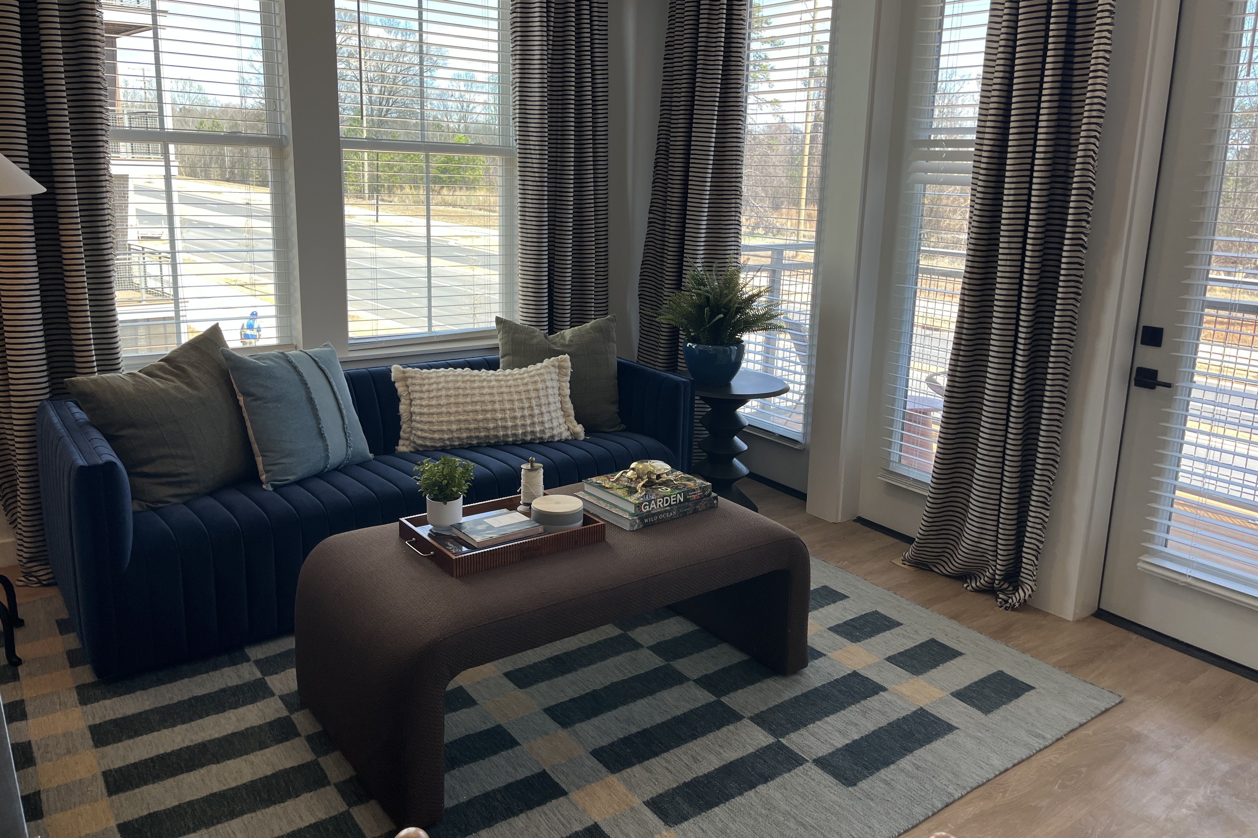 Living room with a navy blue tufted sofa, striped curtains, and a curved brown coffee table on a geometric rug. Green plants sit by large windows overlooking an outdoor deck and bare trees.