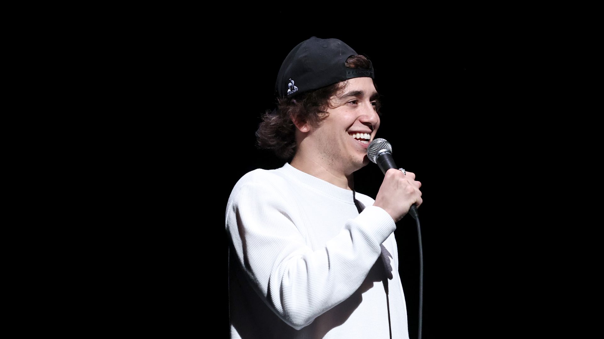 Man wearing a white long sleeve shirt and backwards baseball hat smiling into a microphone. 