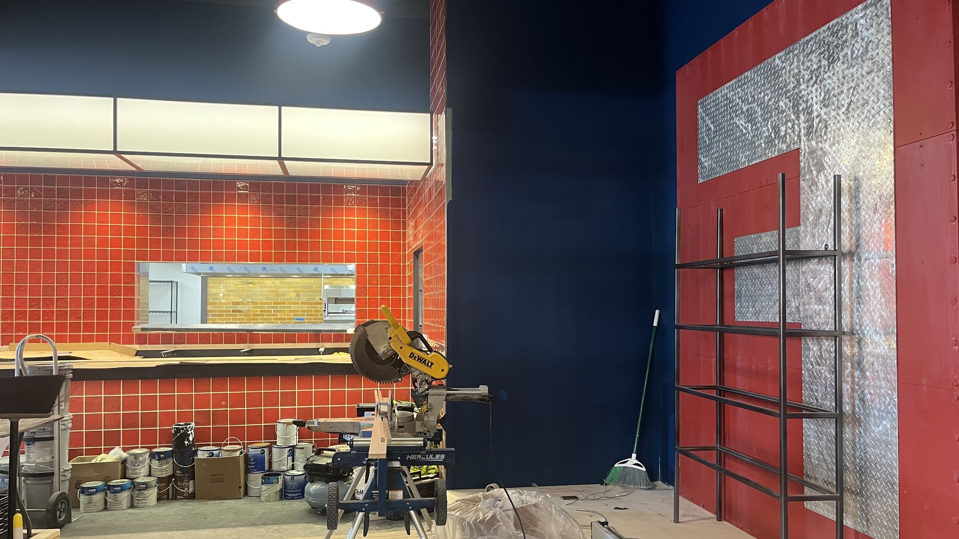 Interior construction site: red tiled wall and blue column, metal diamond-plate panel, scaffolding, a yellow DeWalt miter saw on a stand, tools and paint cans scattered, bright pendant light overhead.