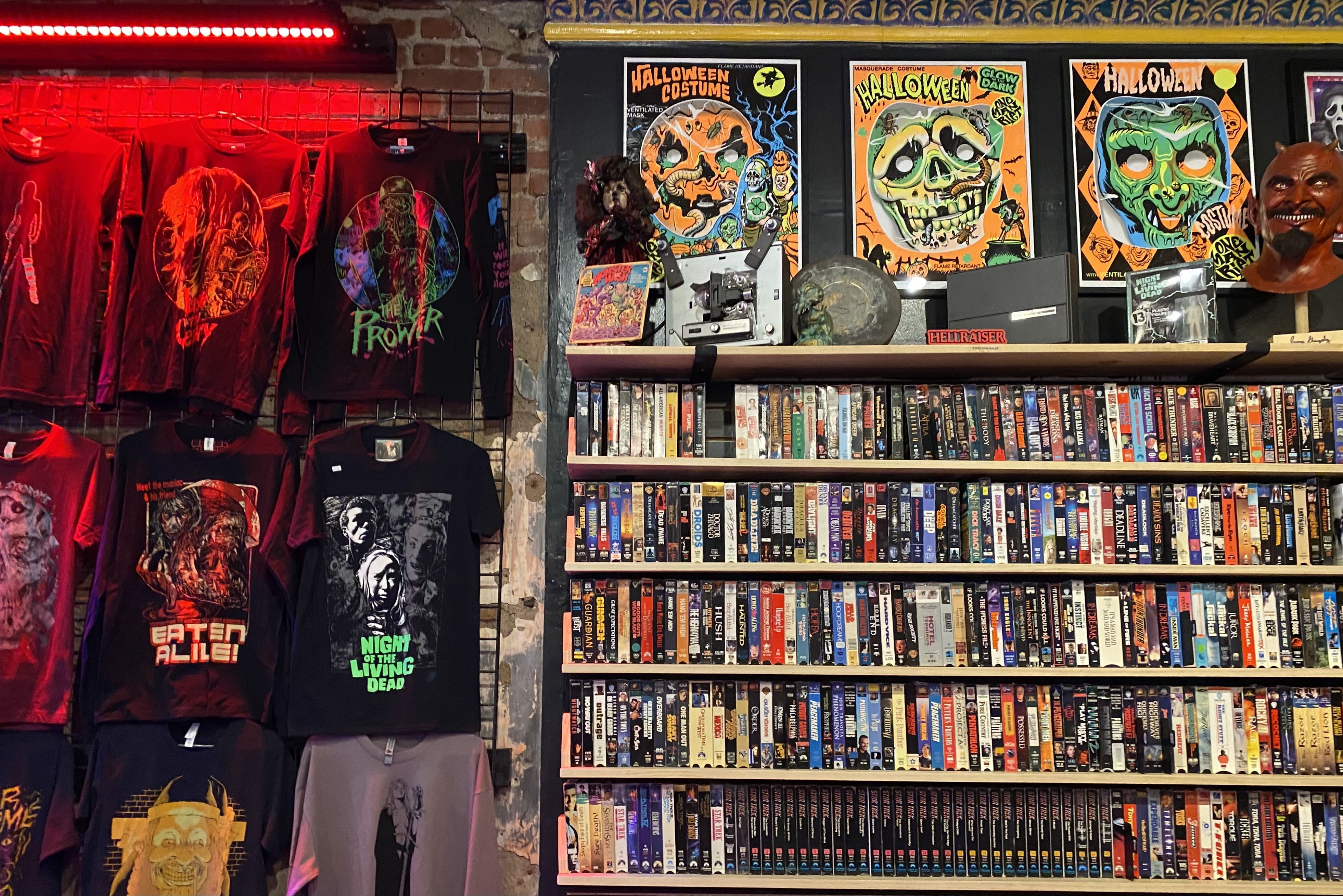 Wall display of horror-themed T-shirts, Halloween posters with skull and pumpkin designs, a shelf with VHS tapes, horror memorabilia, and a red-lit brick wall background.