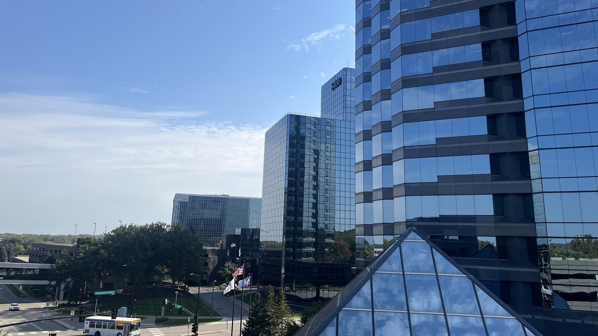 Cityscape showing modern blue glass office buildings reflecting the sky, a pyramid-shaped glass roof in front, trees, a white bus, and street intersection under a clear blue sky.