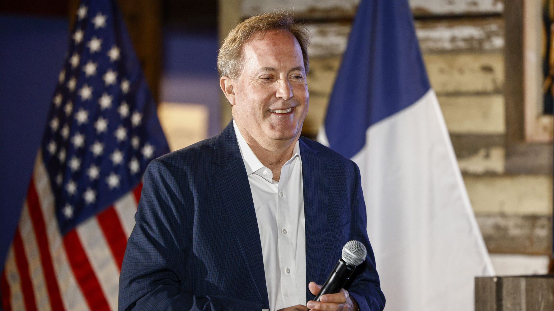 Smiling man in a dark blue suit and white shirt holds a microphone, standing in front of U.S. and French flags at a formal event.