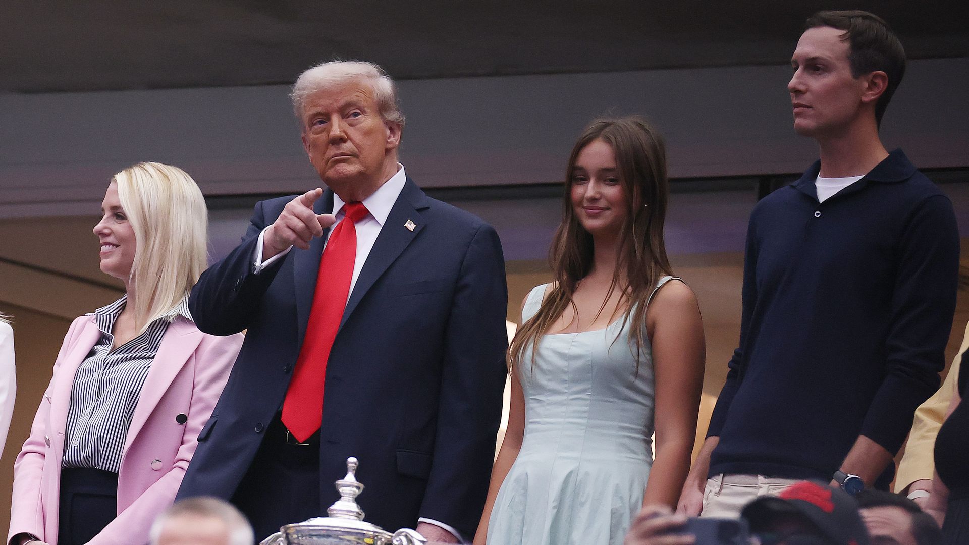 Trump points while attending a tennis match, wearing a dark suit and a red tie. He is flanked by Pam Bondi, who is wearing a pink blazer, a teenage girl in a blue dress.