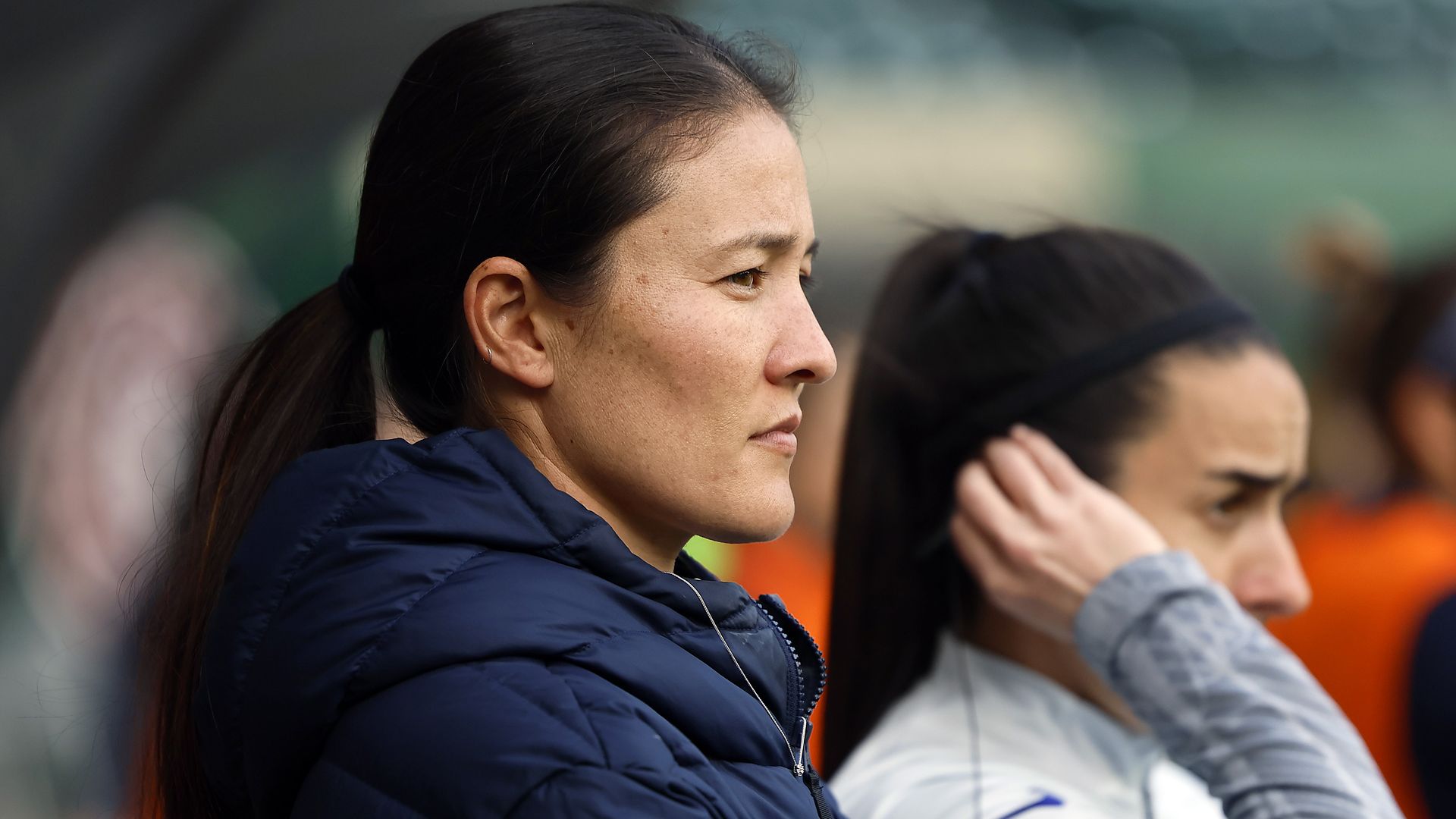 Profile of a woman with dark hair in a ponytail wearing a navy blue jacket, focused and serious, with a blurred woman in white and gray sportswear in the background adjusting her hair.