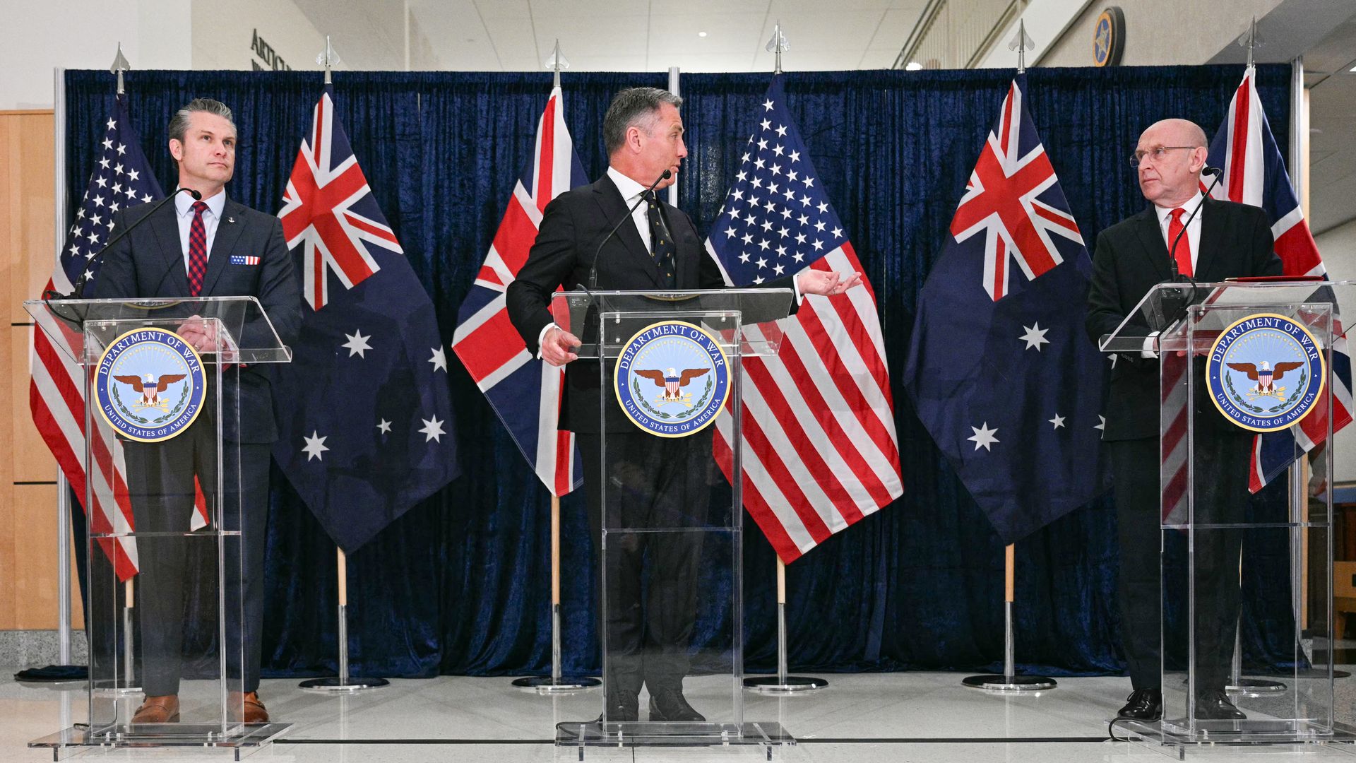 Three men in dark suits stand behind clear lecterns. The American, Australian and British flags hang behind them, against a blue curtain.