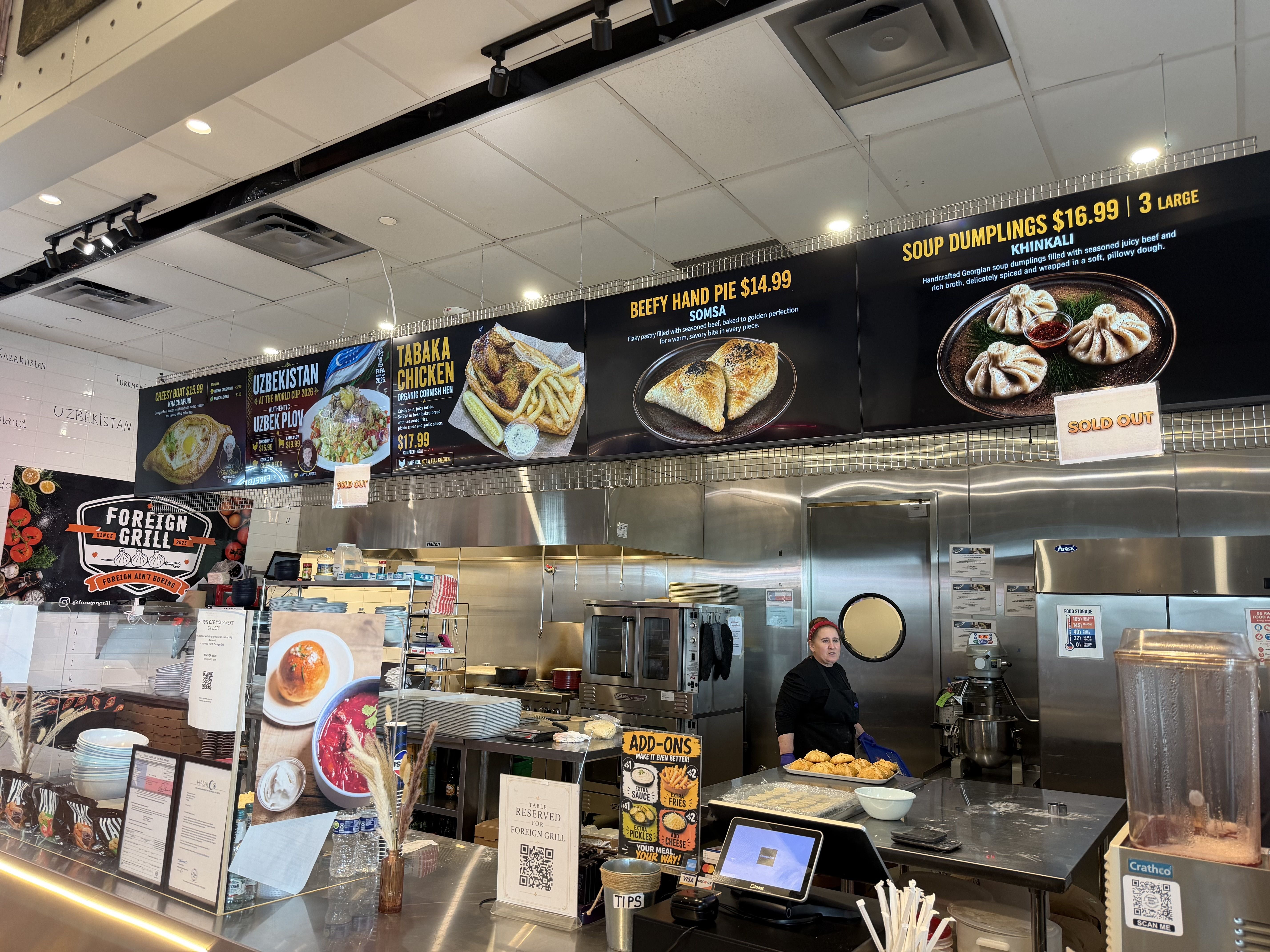 Casual eatery counter with black menu boards showing "SOUP DUMPLINGS $16.99 | 3 LARGE" and "BEEF HAND PIE $14.99". A worker in black stands by a stainless steel kitchen; overhead track lighting.