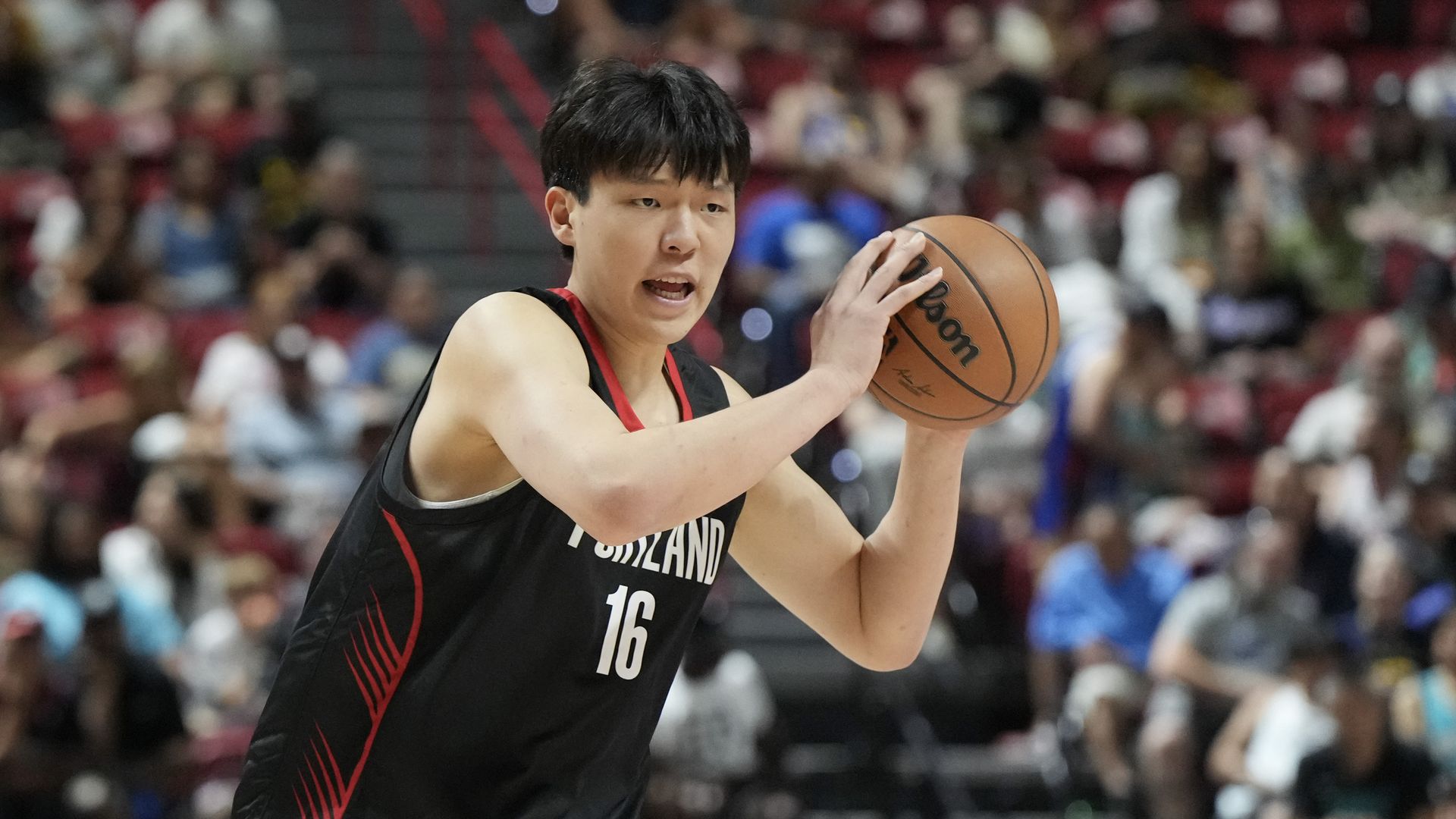 Basketball player in a black Portland jersey with number 16 holds a basketball, preparing to pass or shoot, in an indoor arena with blurred spectators in the background.