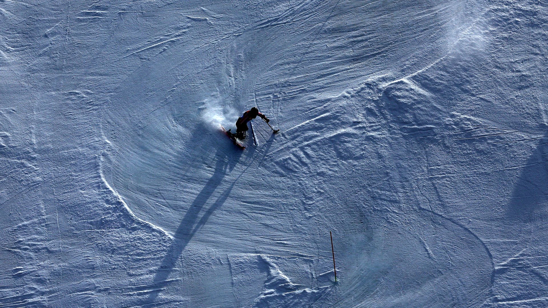 A para skier in dark clothing making a sharp turn on a snowy slope, leaving spray behind. Long shadow stretches left, with ski tracks and a single red ski pole visible in the snow.