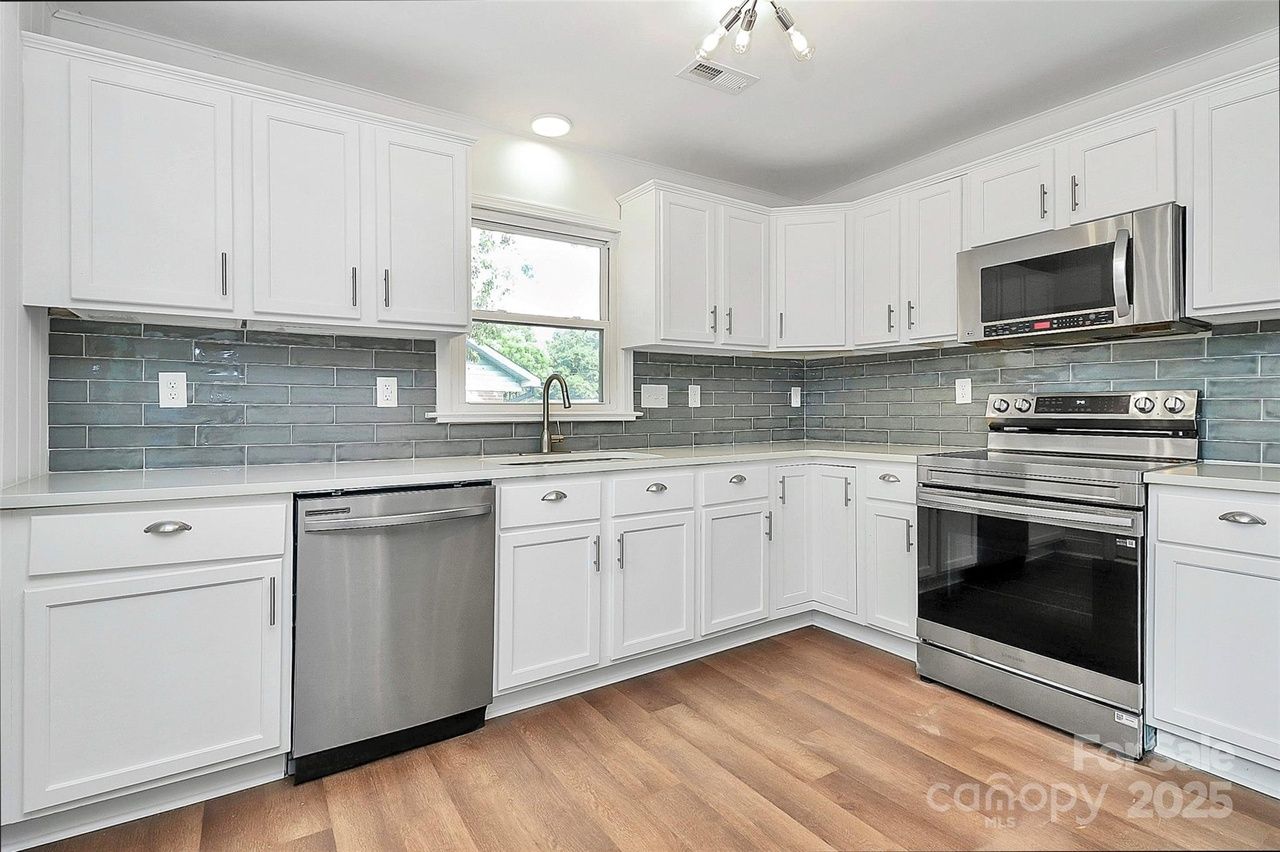 Modern kitchen with white cabinets, gray subway tile backsplash, stainless steel dishwasher, oven, and microwave, wooden floor, and window above the sink.
