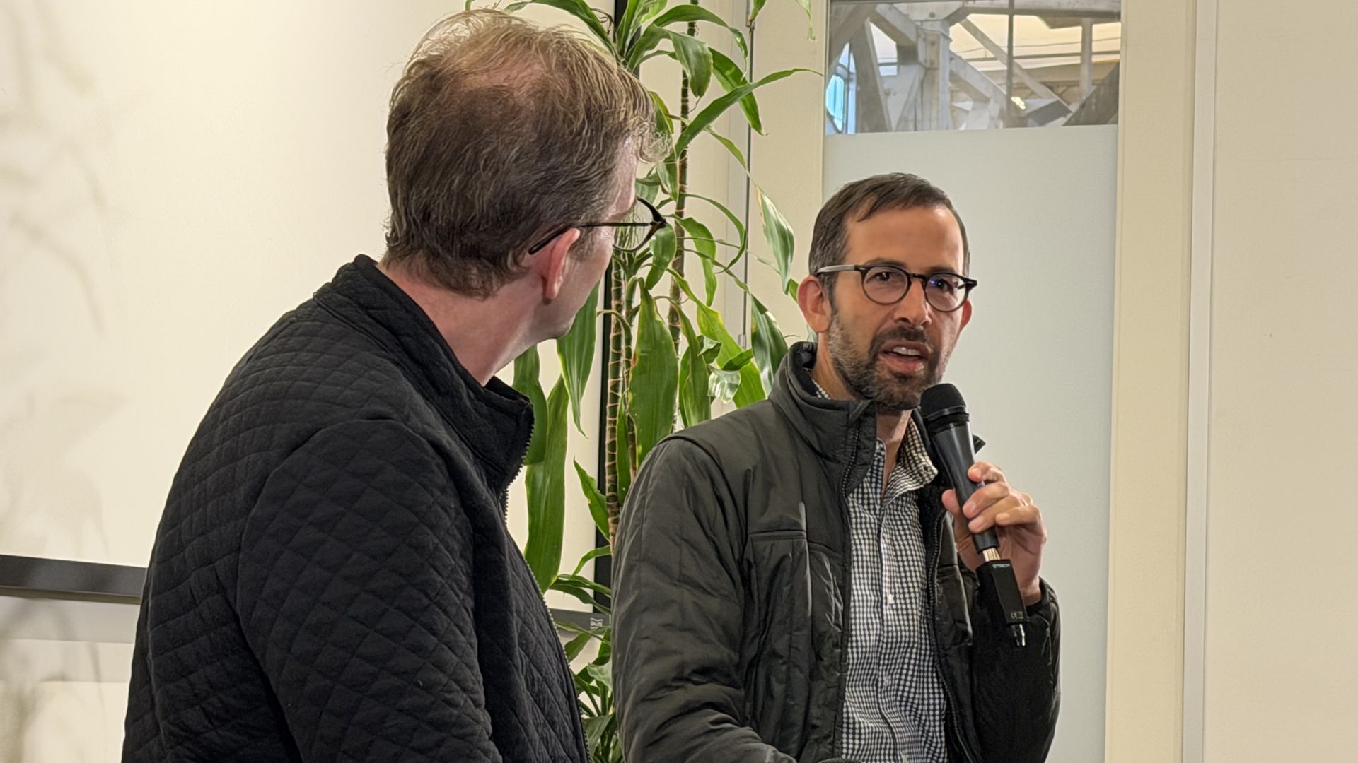 Two men in a casual indoor interview. One man with glasses and a beard speaks into a handheld microphone; the other is seen from the back in a quilted black jacket, beside a tall plant.