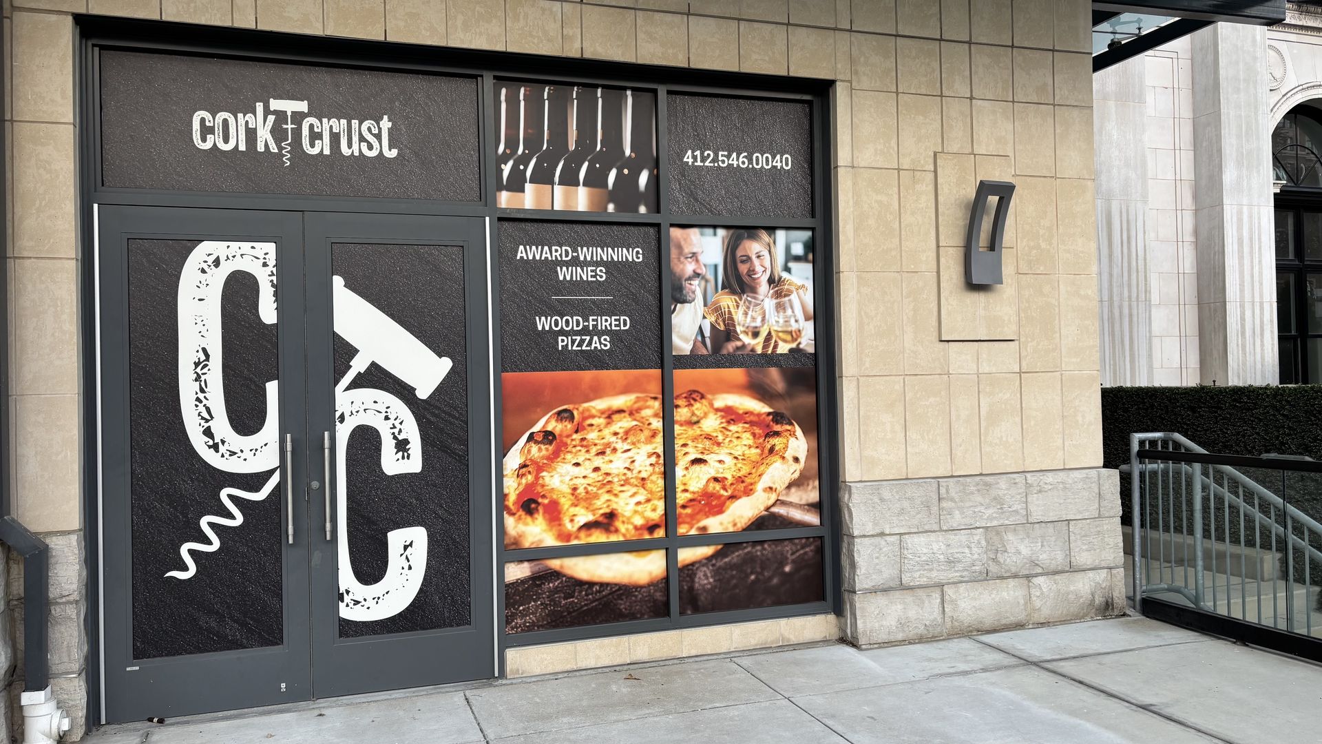 Storefront of Cork Crust showing logo with corkscrew, images of wine bottles, a couple toasting wine glasses, and a wood-fired pizza with text highlighting award-winning wines and pizzas.