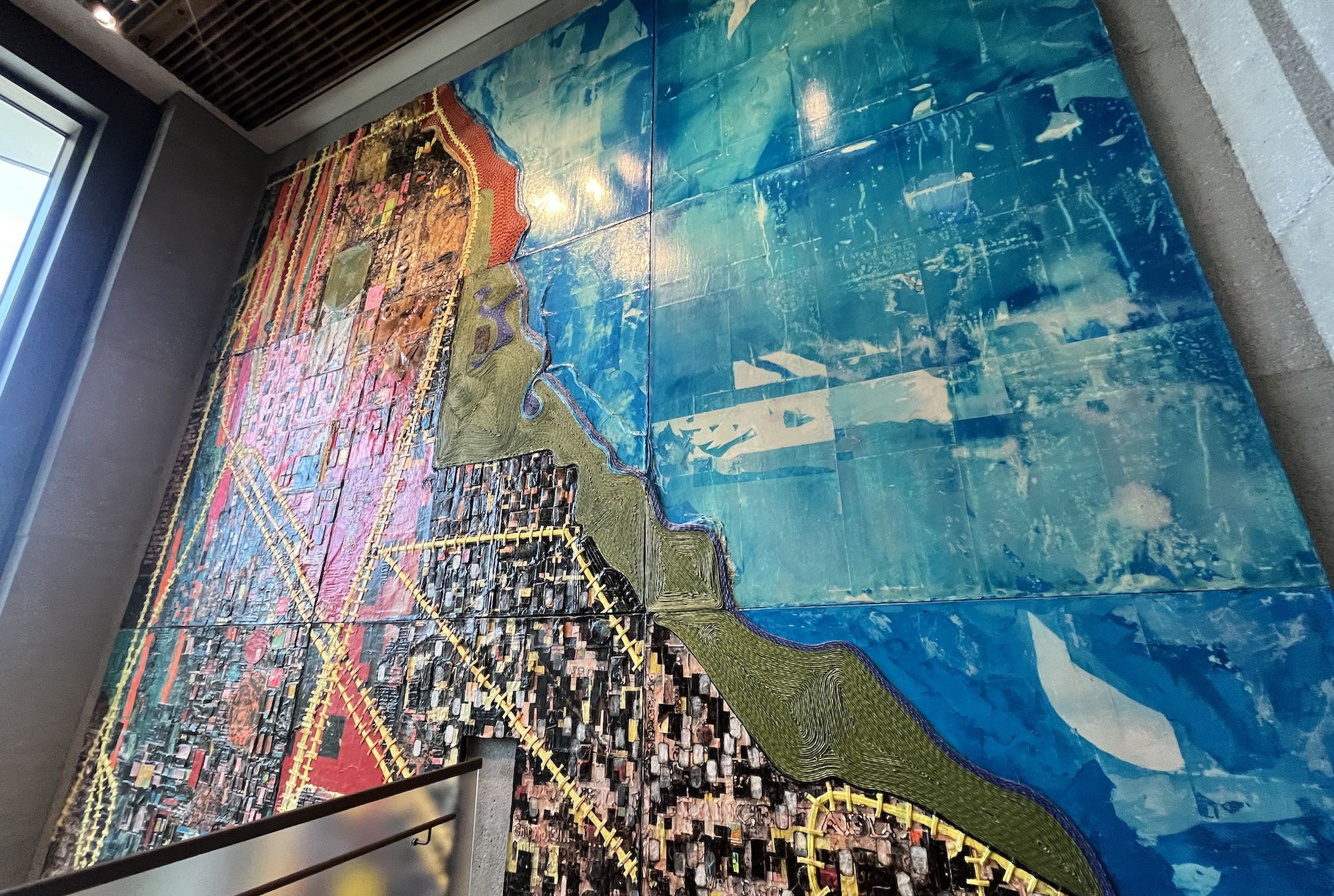 Interior mural: a large colorful mosaic map on a blue tiled wall, with a wavy olive-green border. Left side shows pinks and reds; background blue with light patches. Window and ceiling visible.