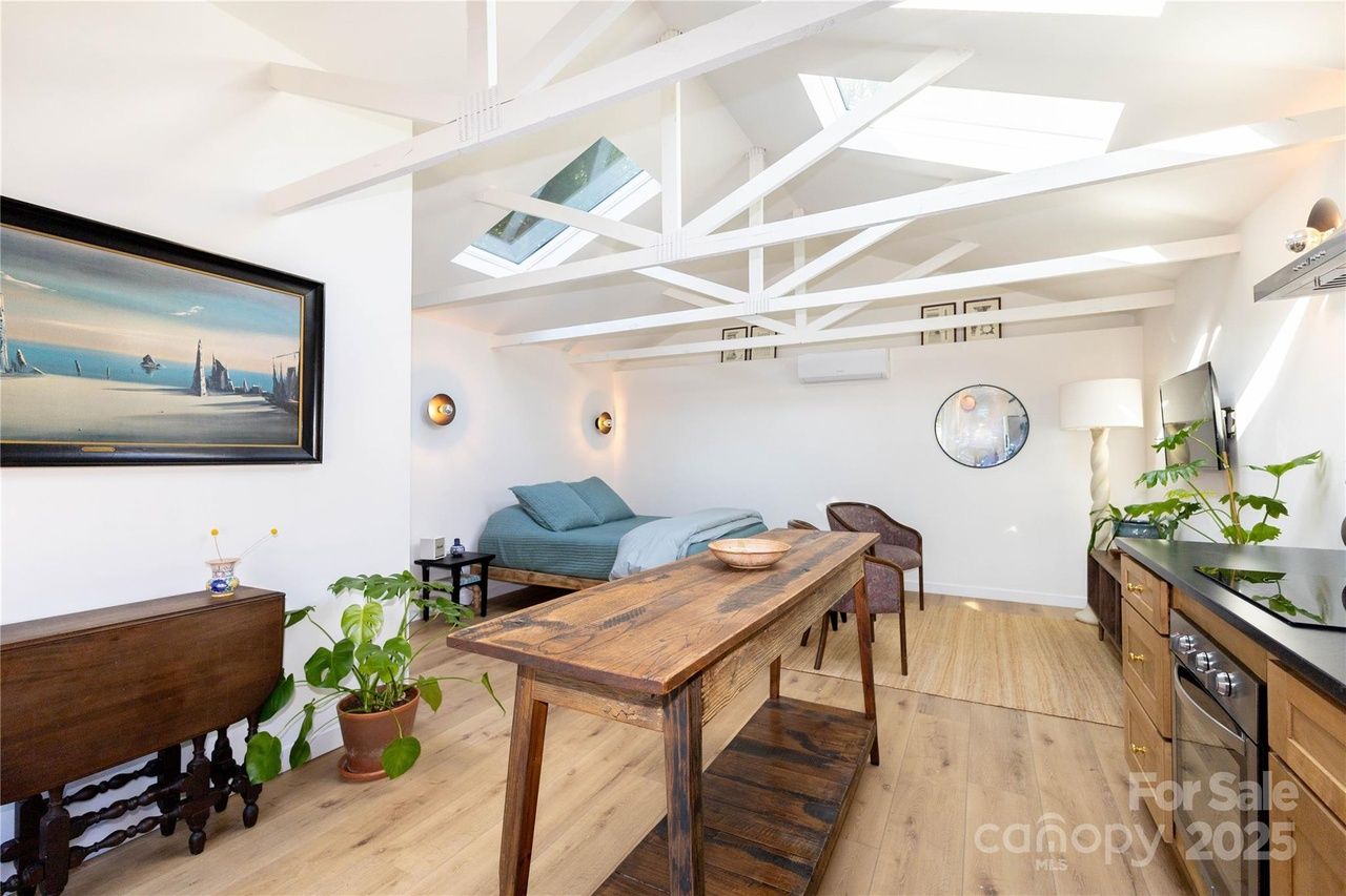 Bright, open studio apartment with white walls, wooden floor and beams, skylights, a bed with teal bedding, wooden dining table, kitchen with stove, plants, and wall art.
