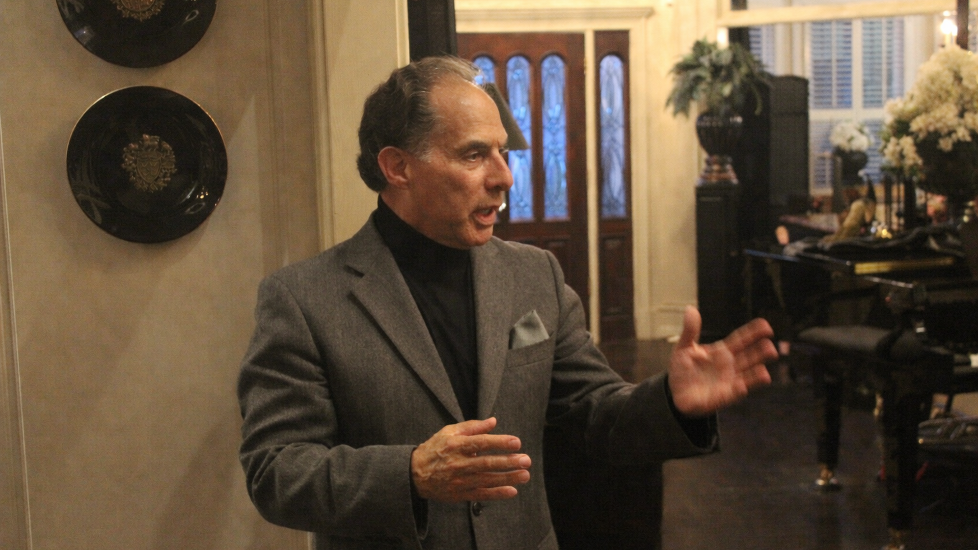 Middle-aged man in a gray blazer and black turtleneck speaks, gesturing with his hands in a luxurious foyer with ornate doors, wall plates, and a piano with flowers in the background.