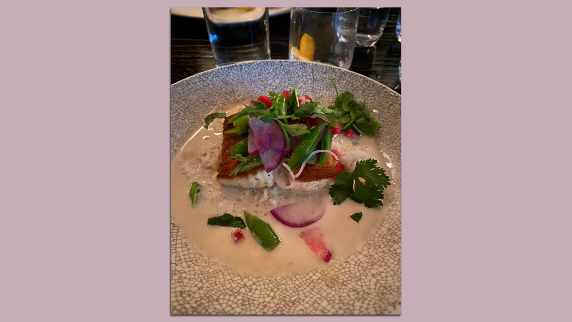 Crusted fish fillet atop creamy white sauce, topped with cilantro, greens, red onion slices, radish, and herbs; served on a gray crackle-pattern plate with glassware in the background.