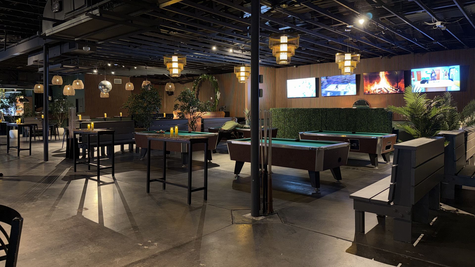 Spacious indoor lounge with pool tables, black benches, warm pendant lantern lights, a row of TVs on a wood-paneled wall, and lush plants under an exposed ceiling.