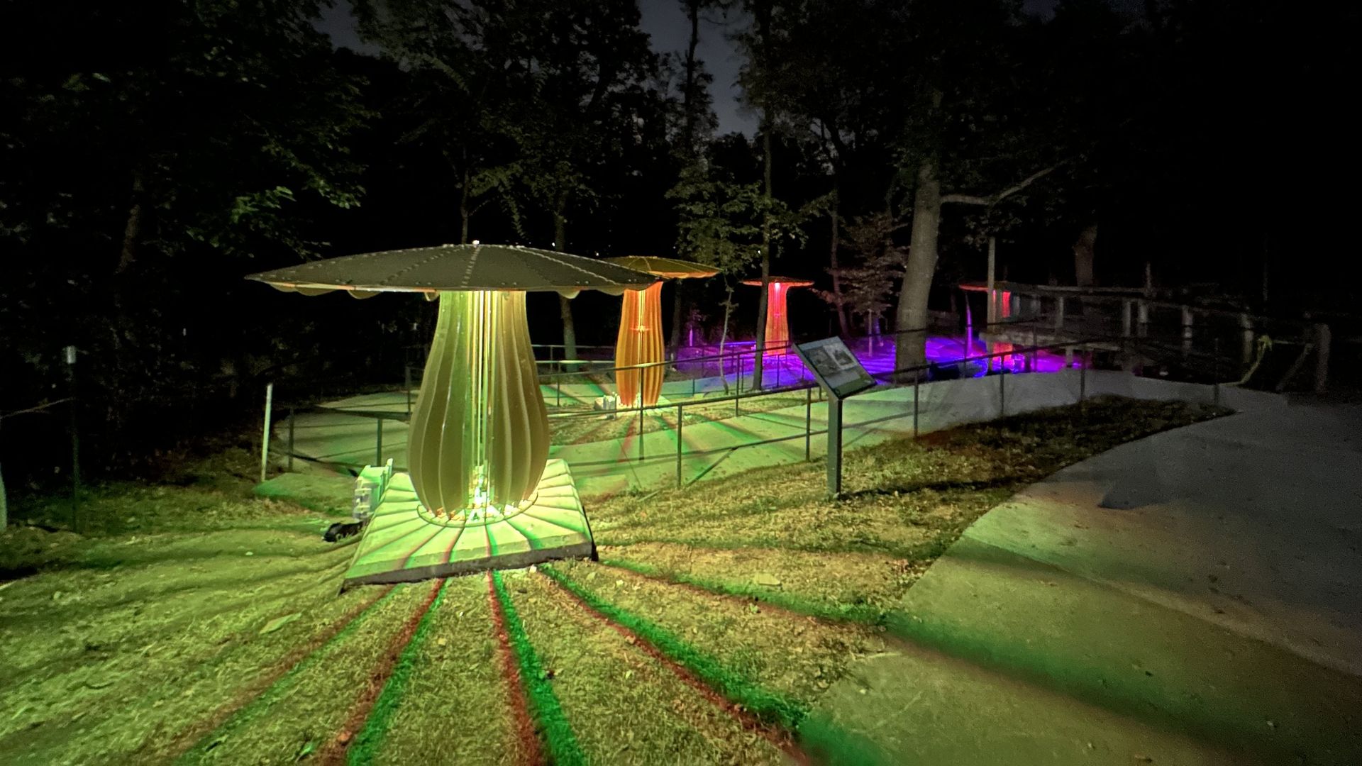 Nighttime outdoor scene with illuminated mushroom-shaped sculptures glowing green, orange, and pink, casting colorful light patterns on the ground and surrounded by trees and a pathway.