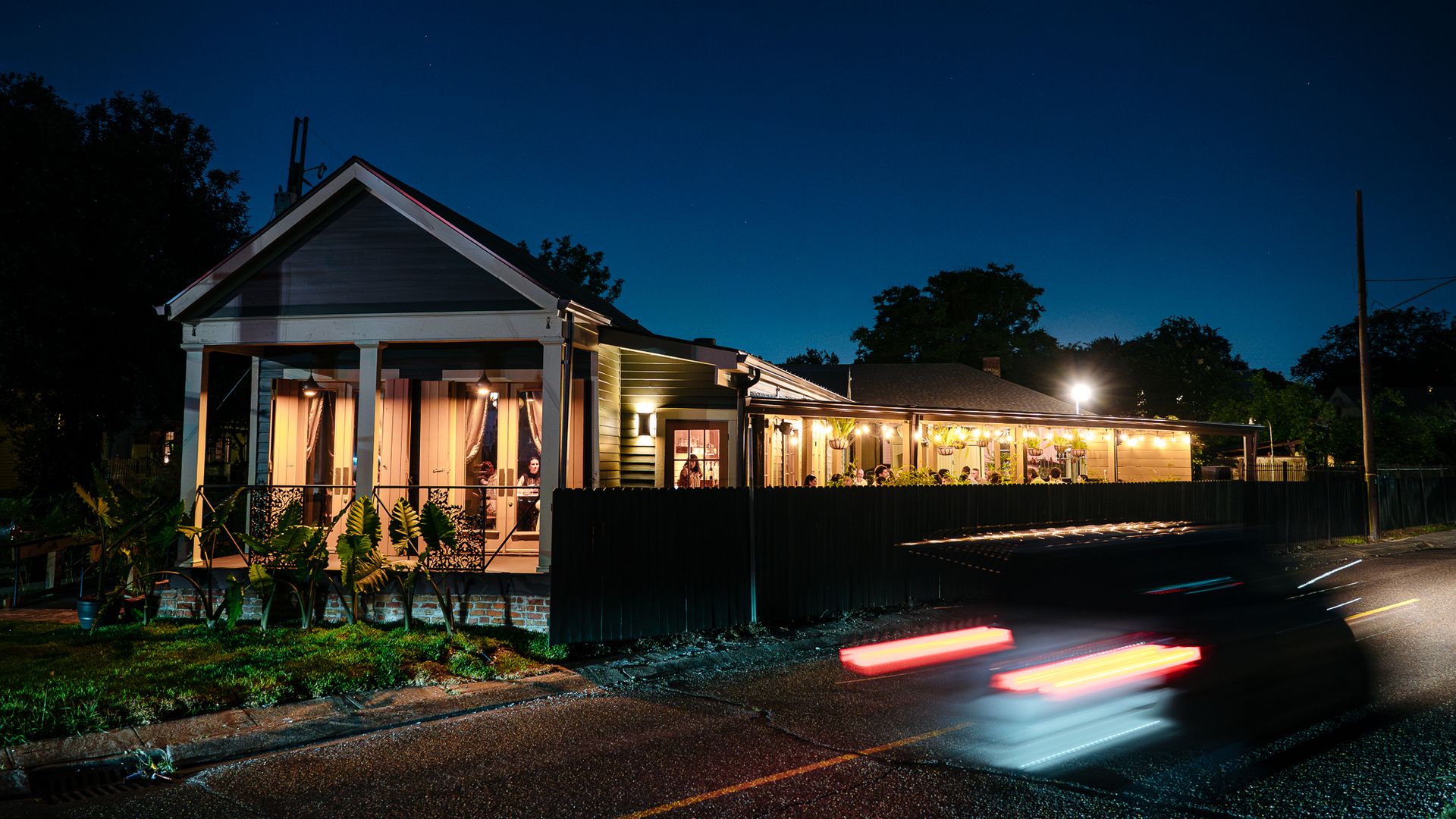 Night view of a house with a gabled roof and warmly lit porch; string lights along the veranda. A black fence, plants in front, and red light trails from passing cars under a deep blue sky.