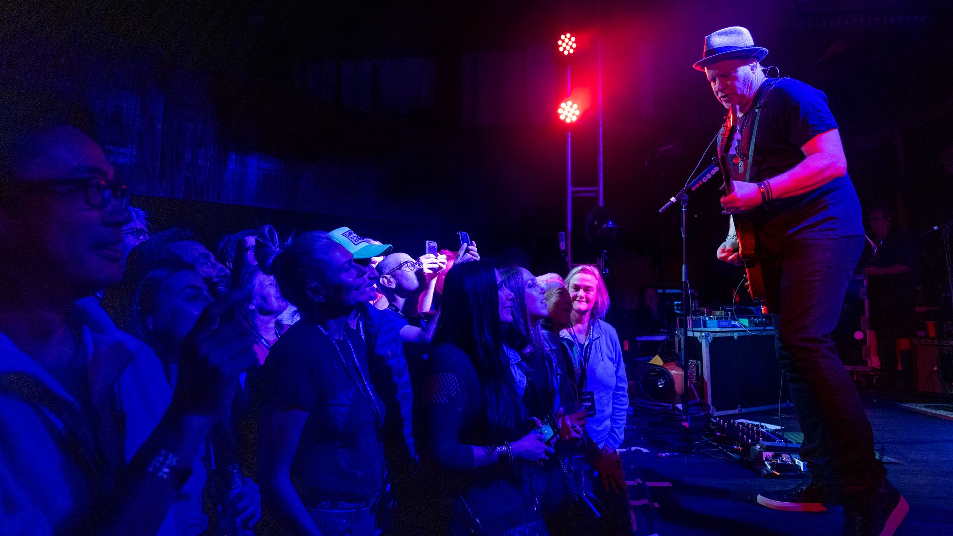 Dan Vickrey holds a guitar while singing into a microphone onstage. At left, a standing-room-only crowd watches the performance.