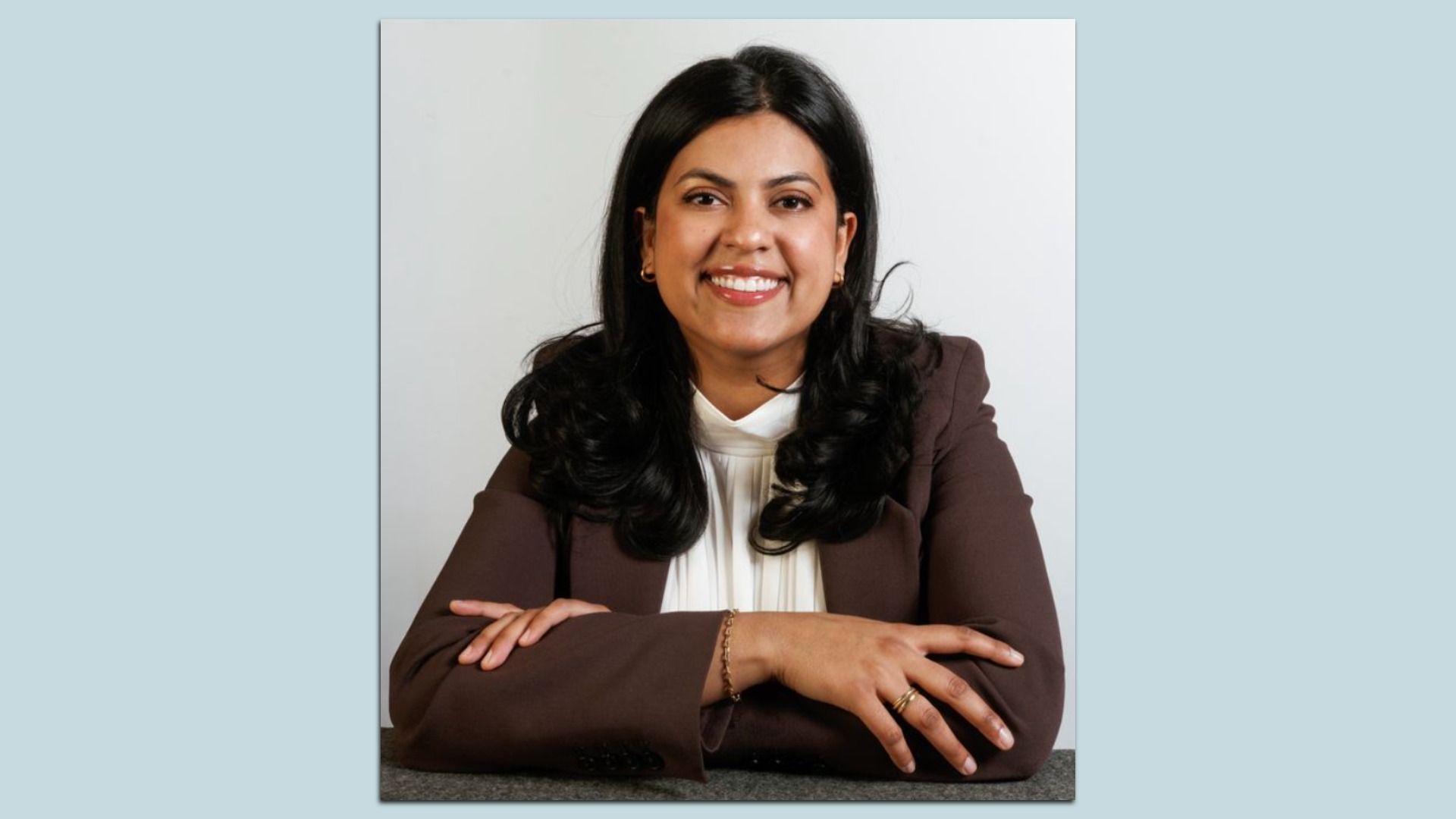 Rini Sampath is photographed sitting with arms crossed on a gray surface against a plain white background.