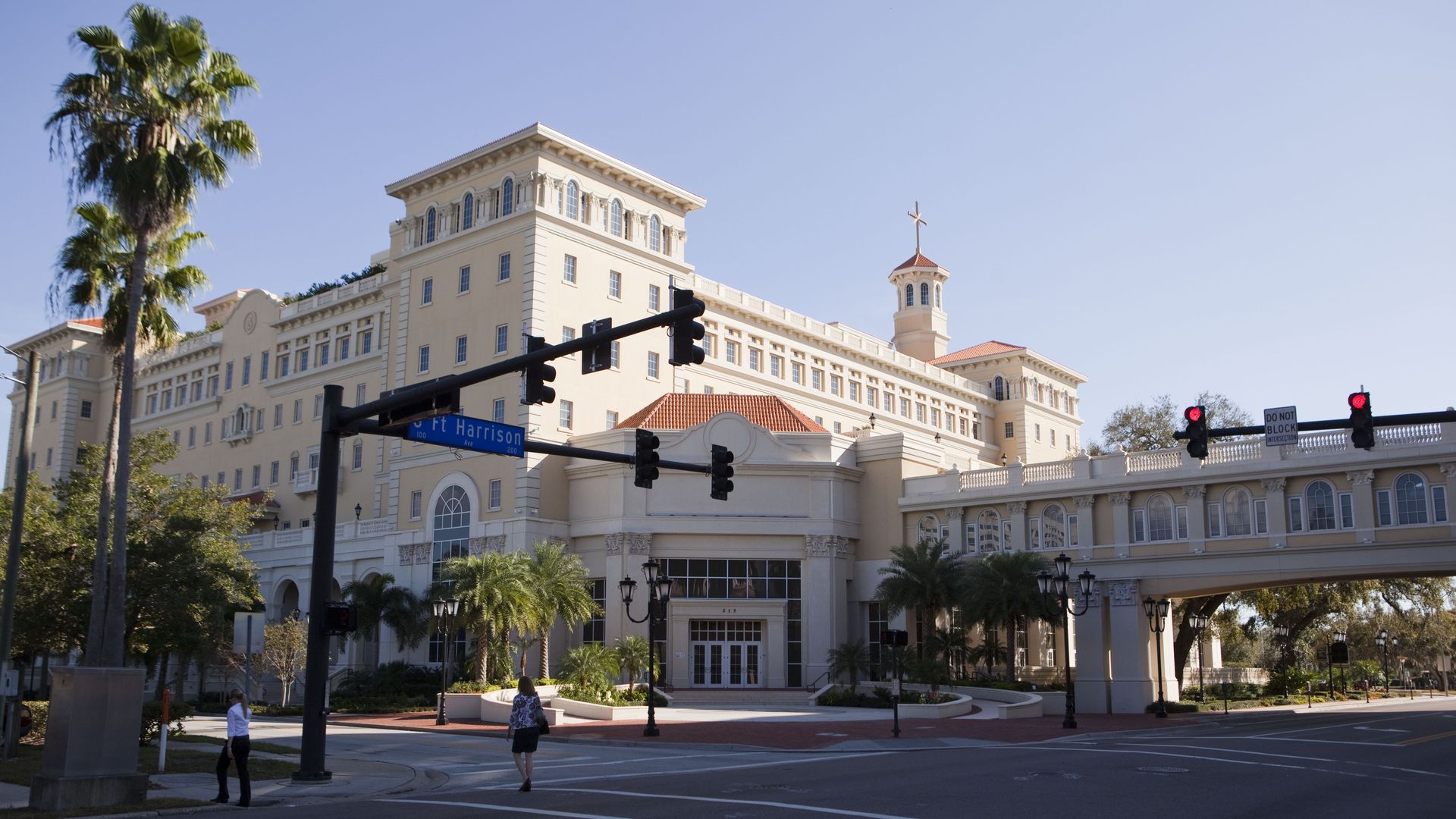 Sunlit beige multi-story building with red-tiled roof and a small tower topped by a cross, flanked by palm trees; clear blue sky, street intersection with traffic signals and pedestrians.