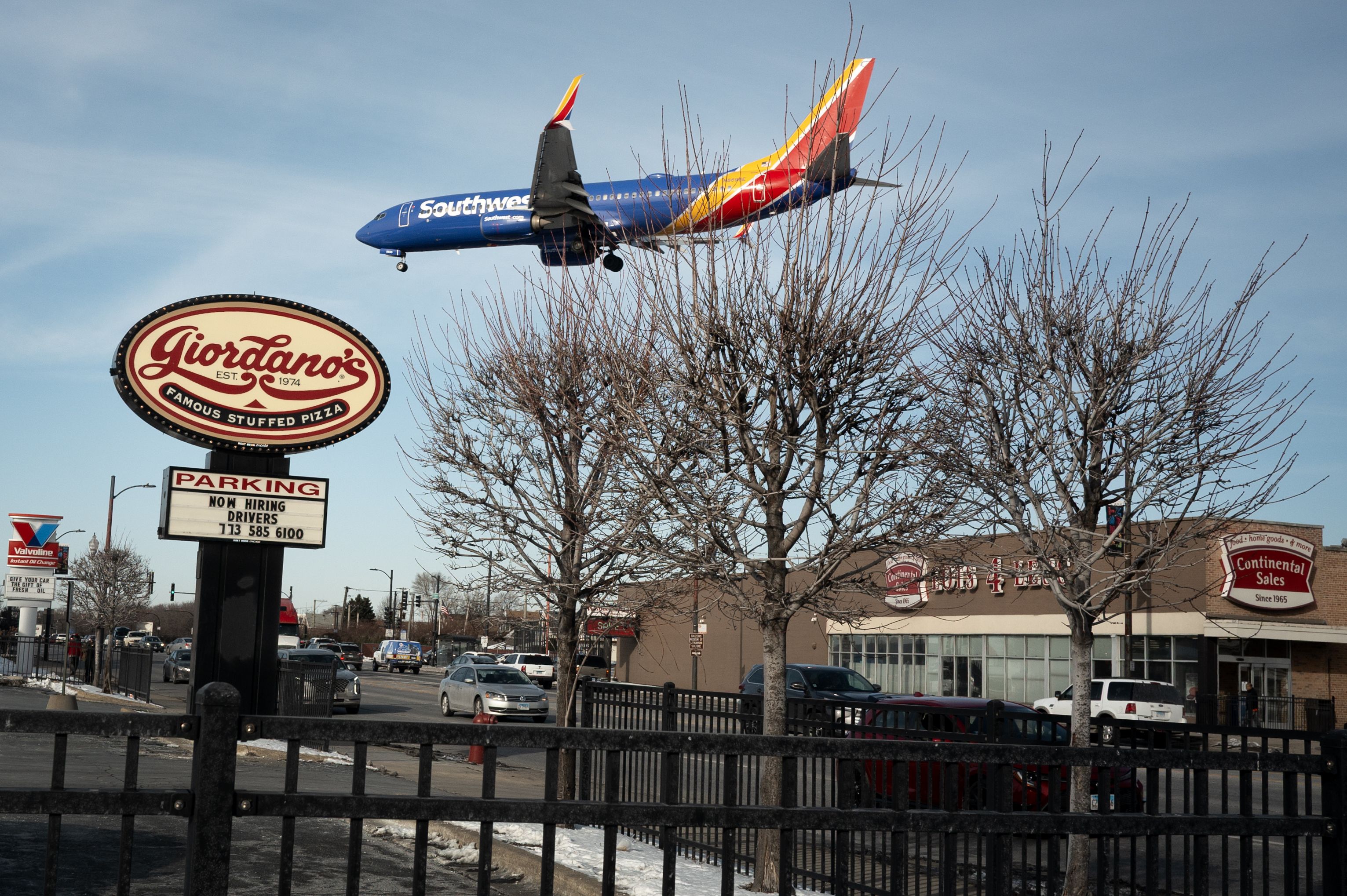 Blue Southwest airplane flying low over a street with leafless trees, cars, and signs including Giordano's pizza and Continental Sales under a clear sky.