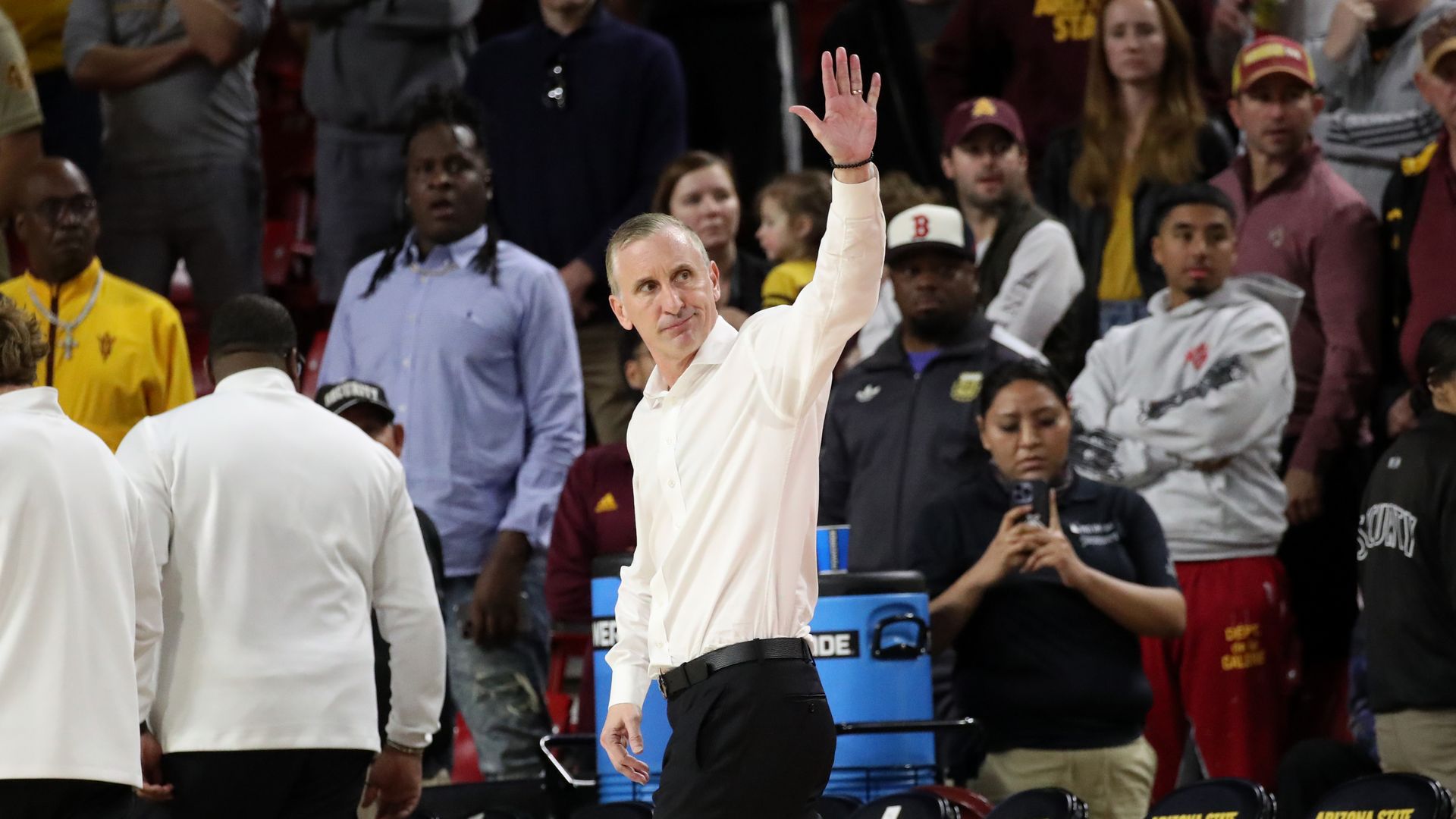 A coach in a white, button-up shirt tucked into black pants looks behind him and waves as he walks off a basketball court, with other people walking away and looking on around him. 