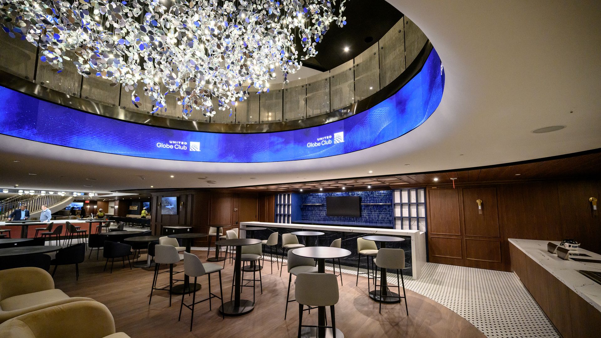 Modern lounge with beige, gray chairs, round tables on wood and tile floors, large metallic chandelier, curved blue digital display reading "United Globe Club".